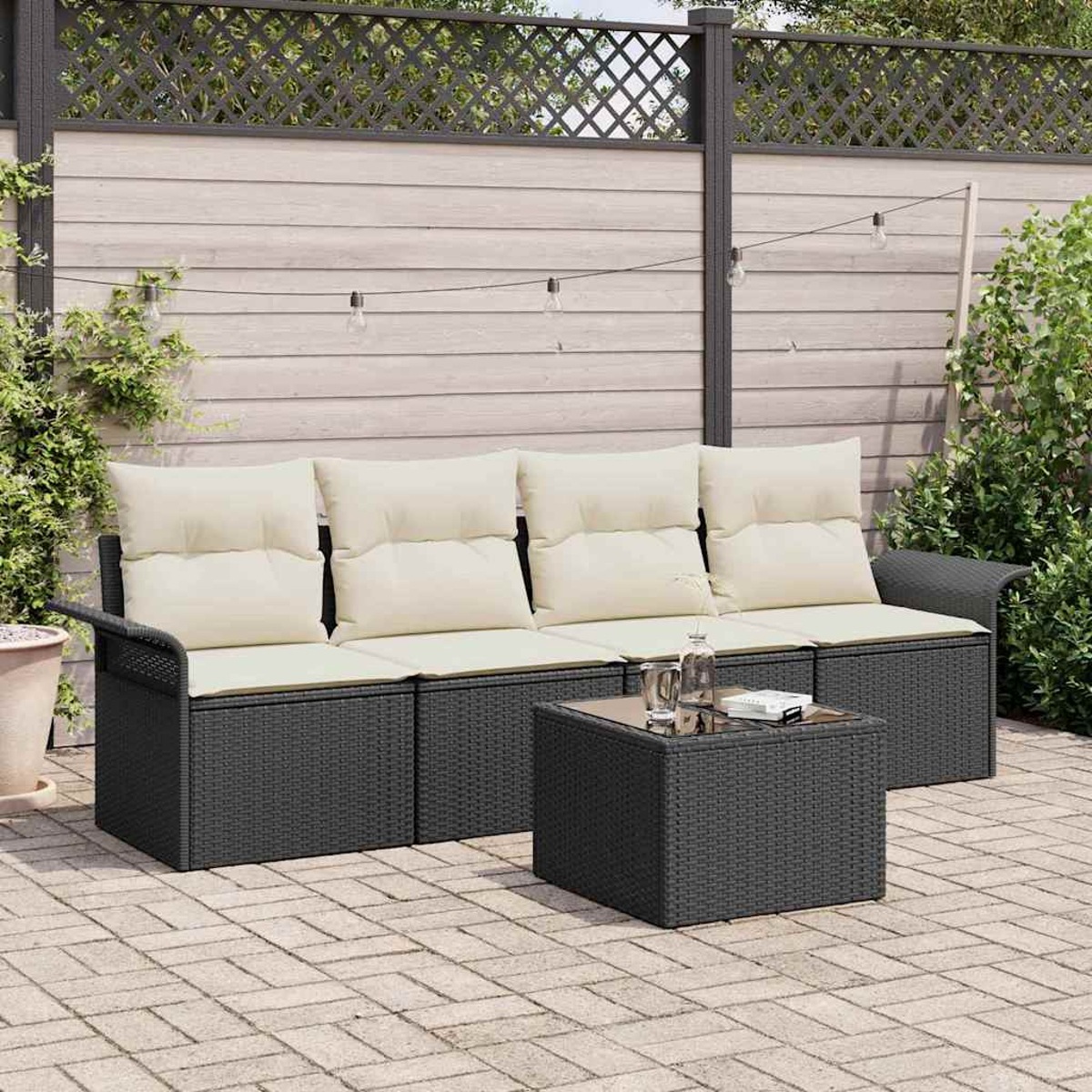 Gartenmöbel-Set: dunkelgraues Korbmöbel-Sofa mit cremefarbenen Kissen, quadratischer Tisch, auf einer Ziegelterrasse. Zaun und Pflanzen im Hintergrund.