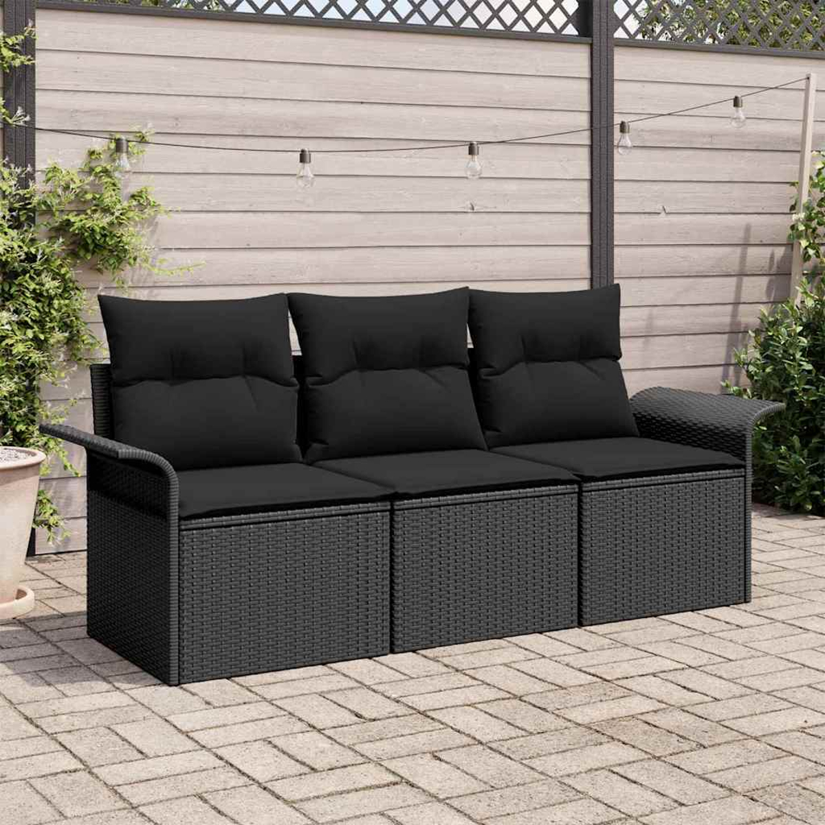 Ein schwarzes Korbmöbel mit schwarzen Kissen steht auf einer gepflasterten Terrasse. Im Hintergrund sind ein Holzzaun und Lichterketten.