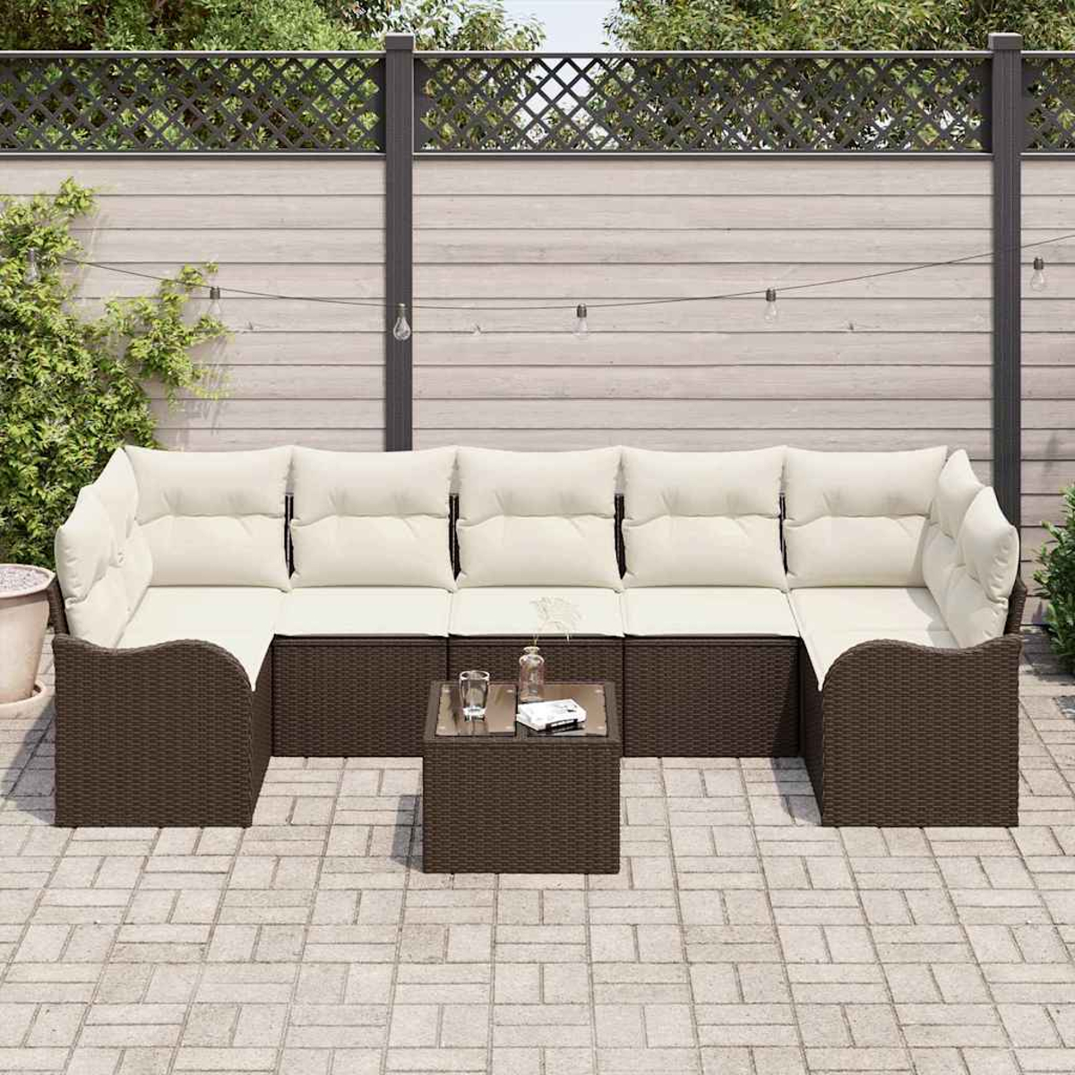 Outdoor-Sofa mit cremefarbenen Kissen und braunem Korbgewebe, kleiner Tisch, auf der Terrasse. Holzzaun-Hintergrund mit Lichterketten.