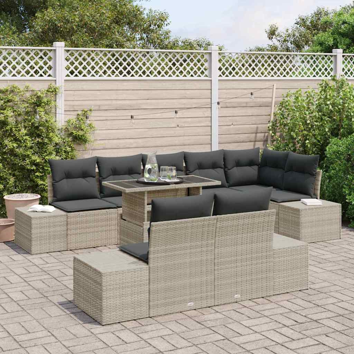 Outdoor-Sitzgruppe mit grauen Kissen. Ein Tisch mit Gläsern und einer Kanne steht in der Mitte. Der Hintergrund hat einen Zaun und Grün.