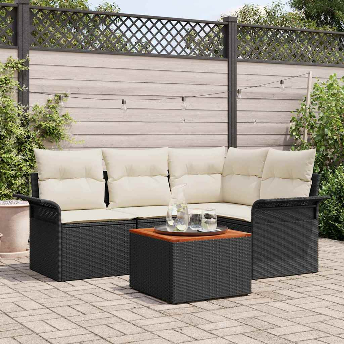 Schwarzes Outdoor-Wicker-Sofa mit cremefarbenen Kissen, ein Holztisch mit Getränken und ein Zaun im Hintergrund.