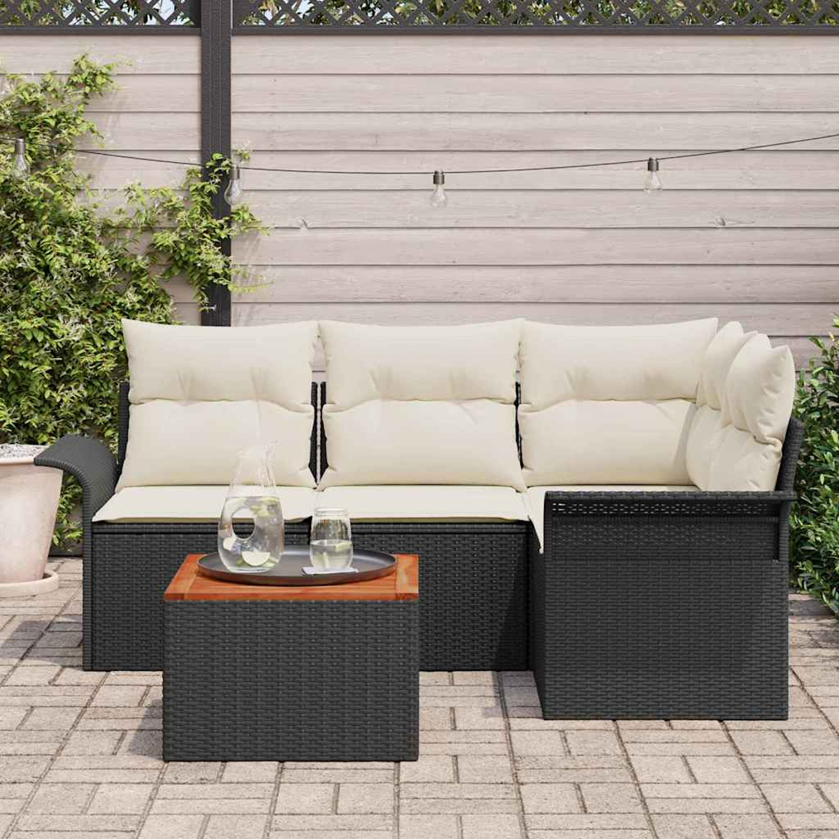 Outdoor-Sitzbereich. Schwarzes Korbmöbel mit cremefarbenen Kissen. Ein kleiner Tisch mit Tablett und Getränken. Holzzaun und Pflanzen im Hintergrund.
