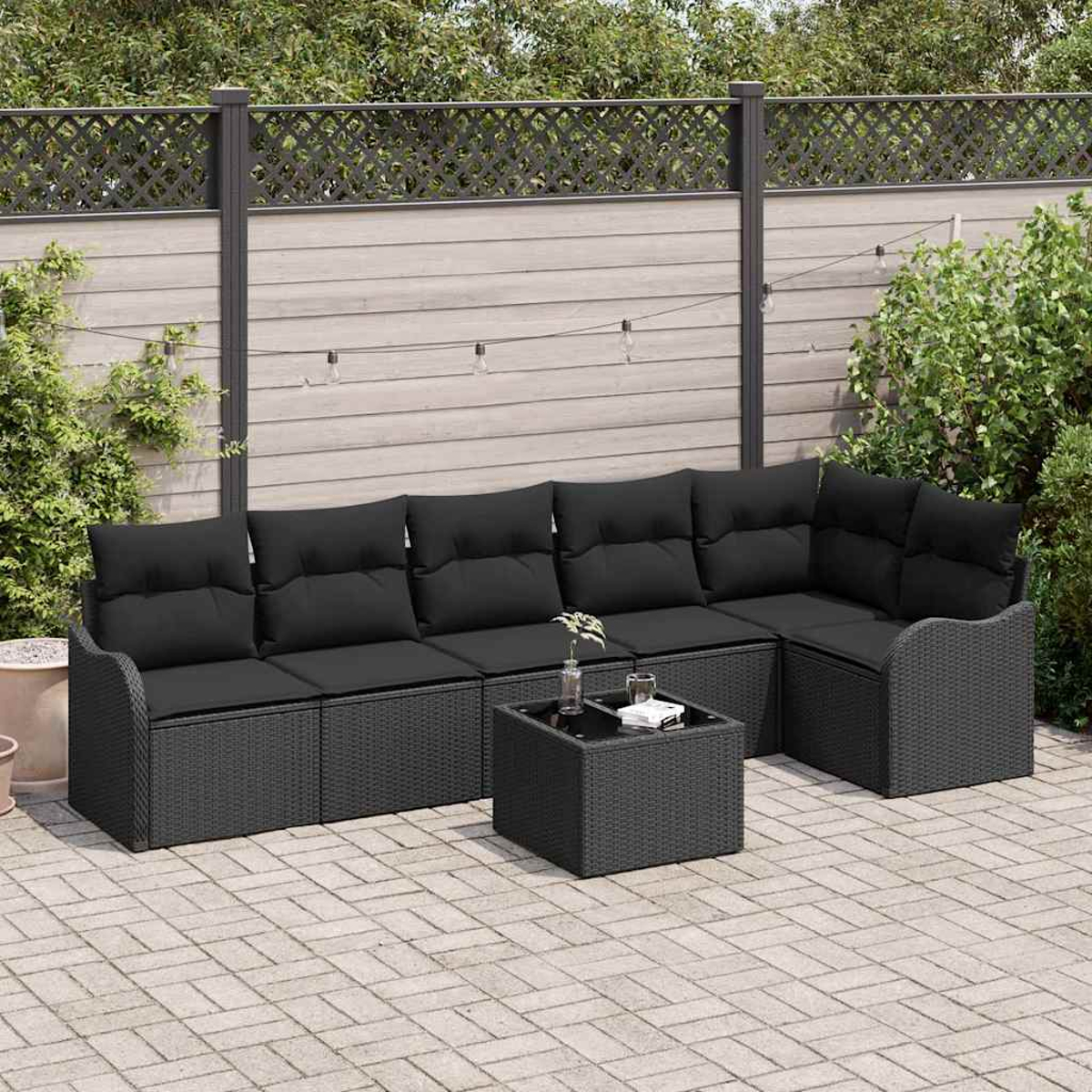 Outdoor-Ecksofa mit schwarzen Kissen und einem Couchtisch auf einer Backstein-Terrasse. Im Hintergrund ein Zaun und Grün.