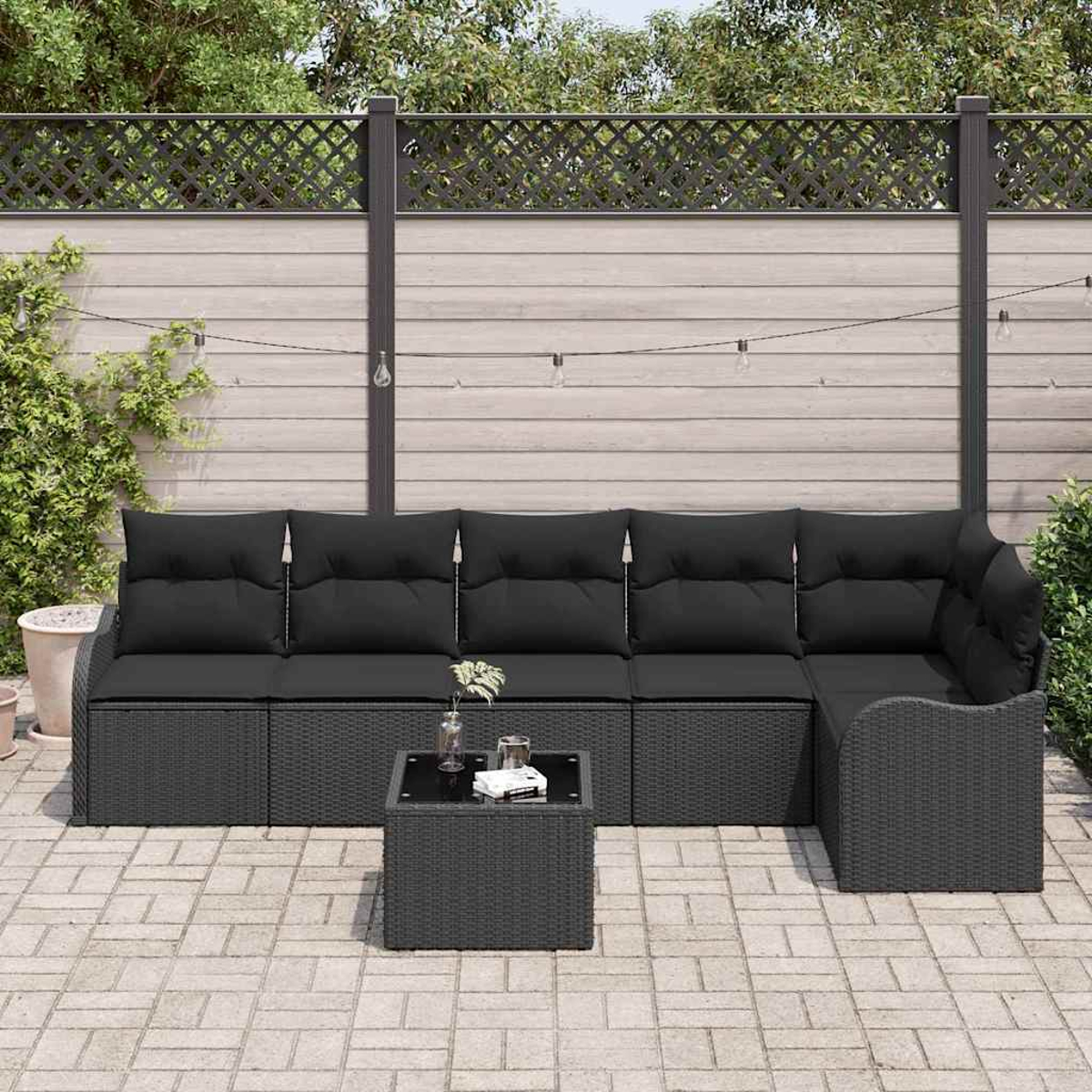 Ein schwarzes Rattan-Sofa mit schwarzen Kissen und ein quadratischer Couchtisch stehen auf einer Terrasse. Ein Holzzaun und Grünzeug sind im Hintergrund.
