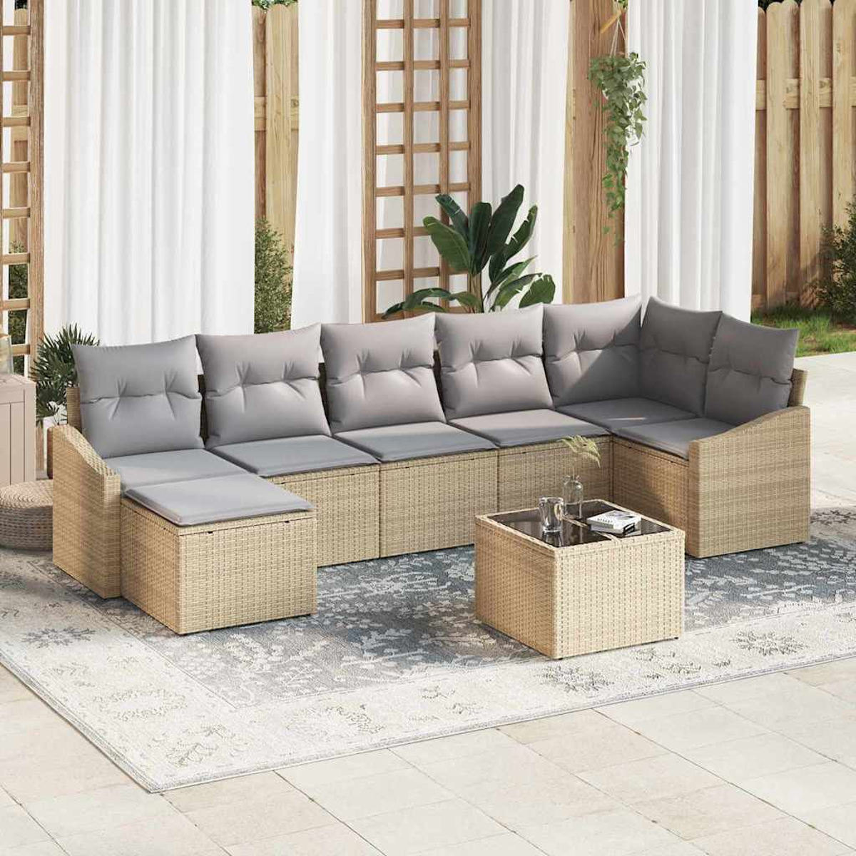 Geflochtenes Ecksofa mit grauen Kissen, Couchtisch und einem gemusterten Teppich auf einer Terrasse. Weiße Vorhänge und Holzzaun im Hintergrund.