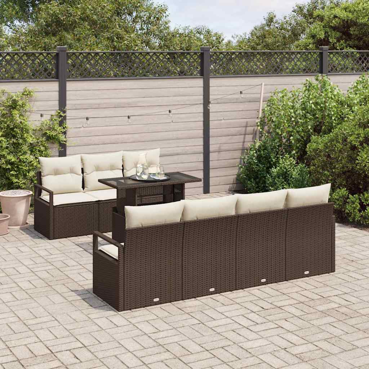 Garten-Terrassenset: braune Korbsessel mit cremefarbenen Kissen, ein kleiner Tisch und ein gepflasterter Boden vor einem Zaun und Grün.