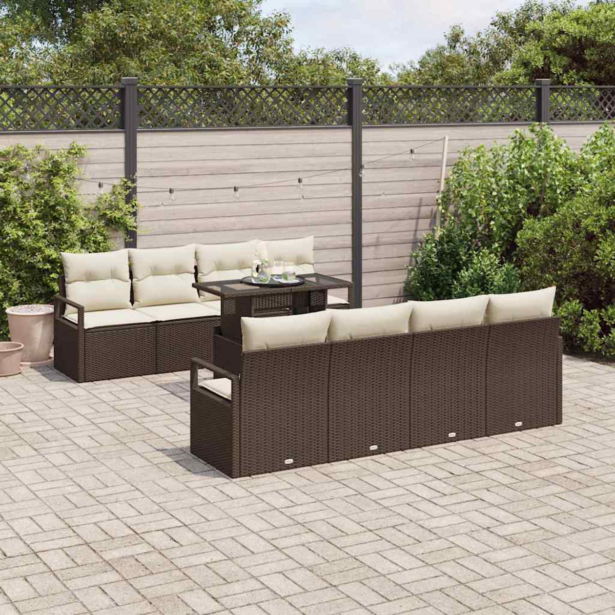 Garten-Set mit braunen Korbmöbeln und cremefarbenen Kissen auf einer Ziegelterrasse. Ein Tisch, Pflanzen und ein Zaun befinden sich im Hintergrund.