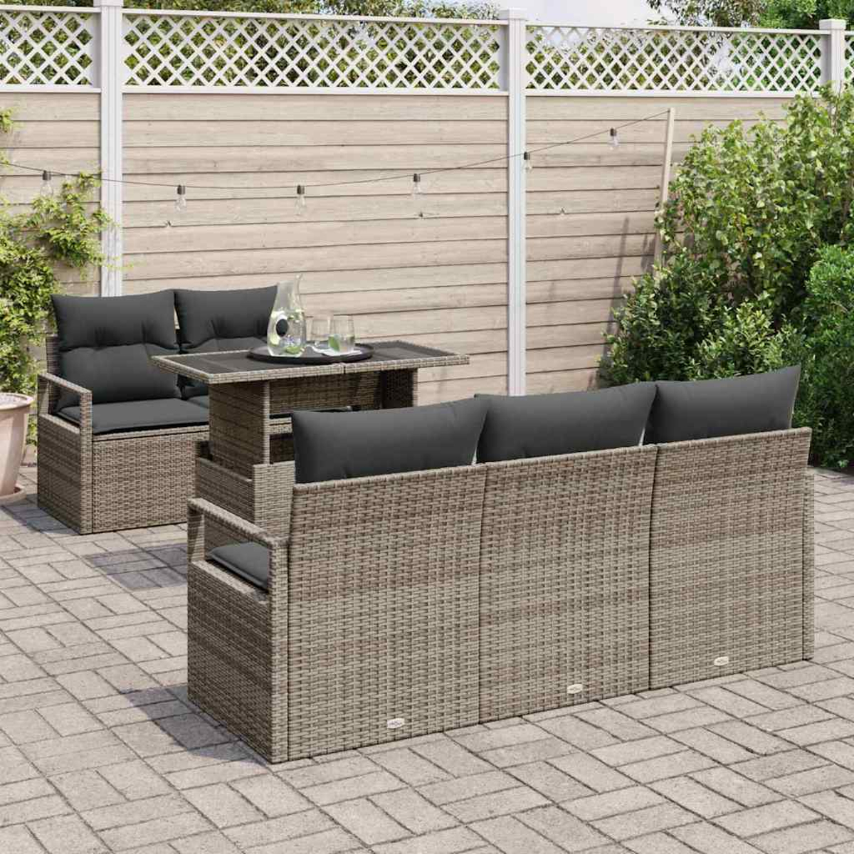 Terrassengarnitur: graues Korbmöbel-Sofa, zwei Stühle und Tisch mit dunklen Kissen. Hintergrund: Holzzaun und Grün.
