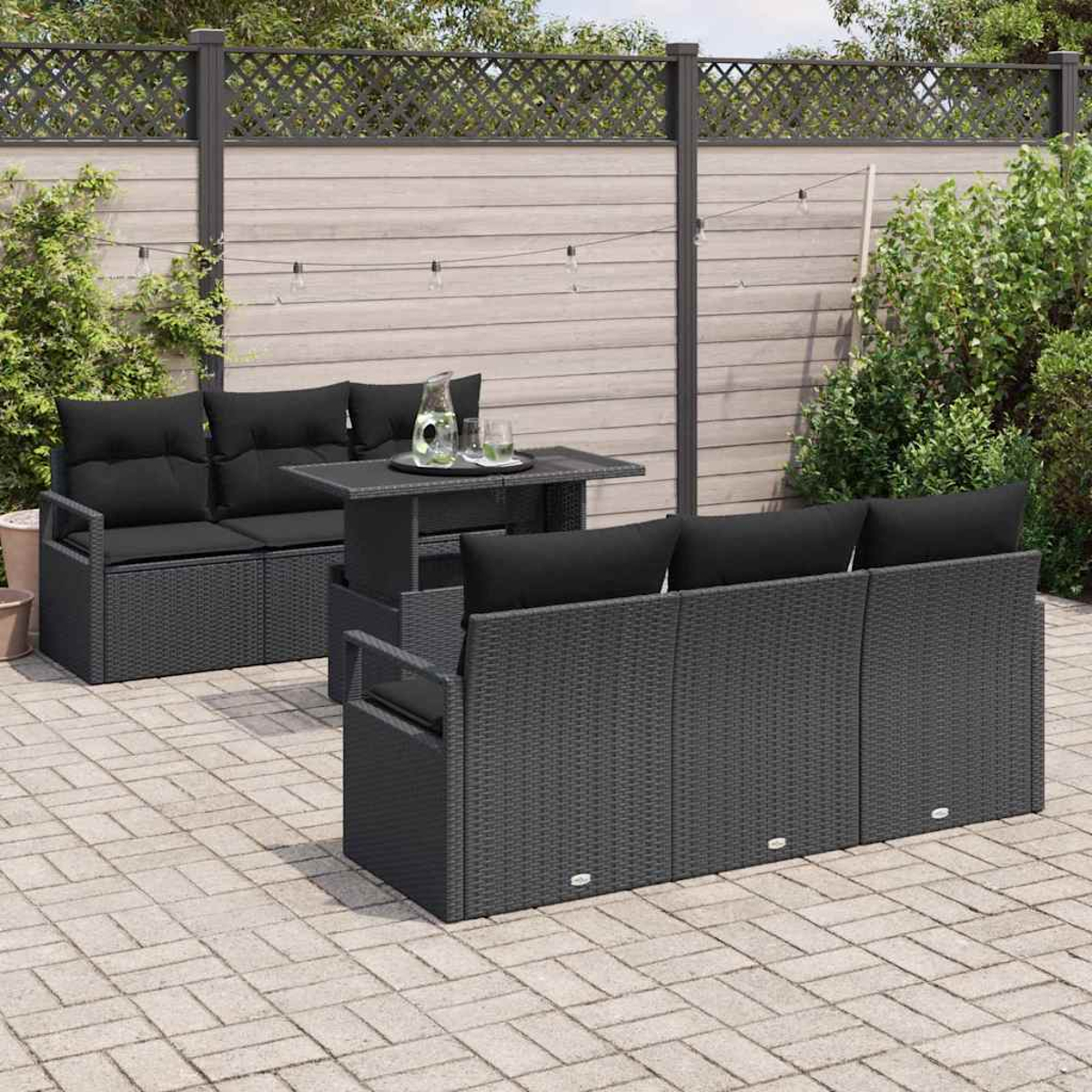 Terrassenset im Freien. Dunkelgraues Korbmöbel-Sofa, Tisch und Stühle mit schwarzen Kissen auf Steinterrasse, Holzzaun im Hintergrund.