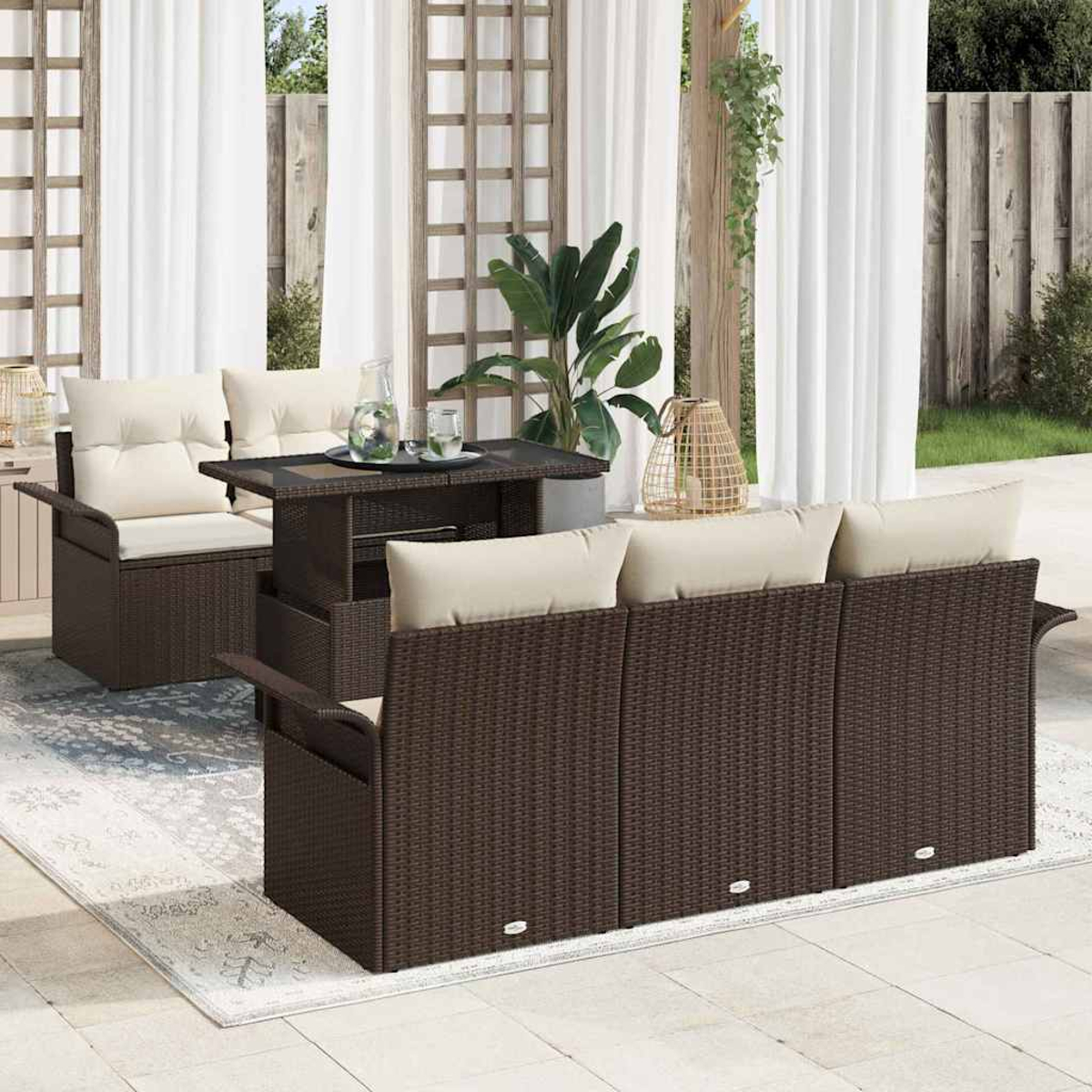 Juego de patio al aire libre con muebles de mimbre marrón, cojines crema, una mesa y cortinas blancas. Plantas en el fondo.