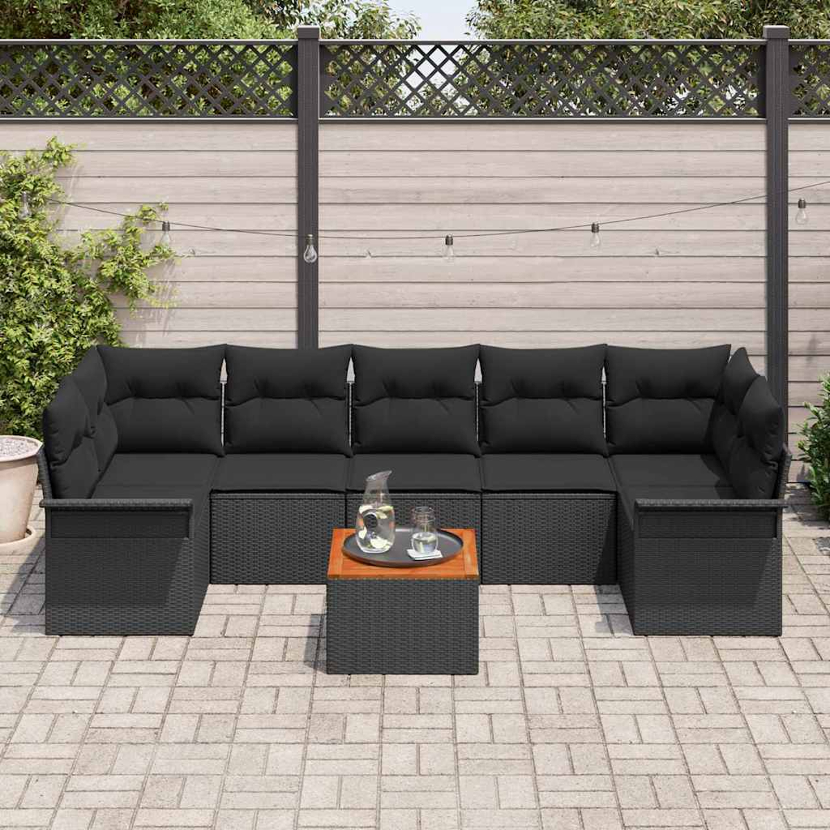 Ein schwarzes Korbsessel-Sofa mit schwarzen Kissen steht auf einer Ziegelsteinterrasse. Ein kleiner Holztisch und ein Zaun sind im Hintergrund.
