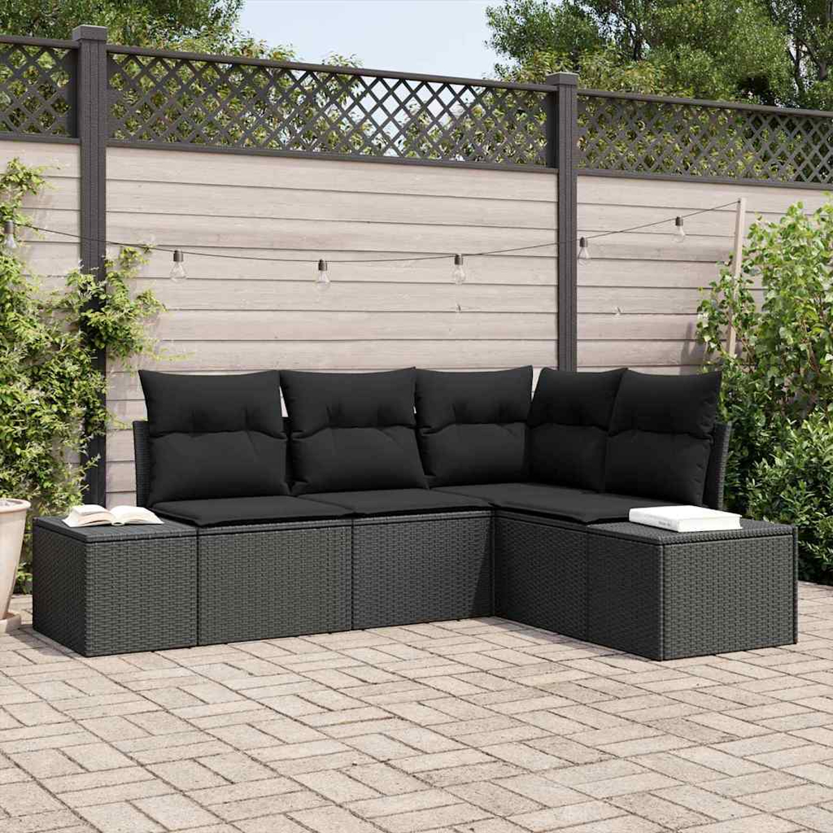 Schwarzes Ecksofa aus Korbgeflecht im Freien mit schwarzen Kissen auf einer Backstein-Terrasse. Ein Buch liegt auf einem Beistelltisch. Im Hintergrund ein Holzzaun.