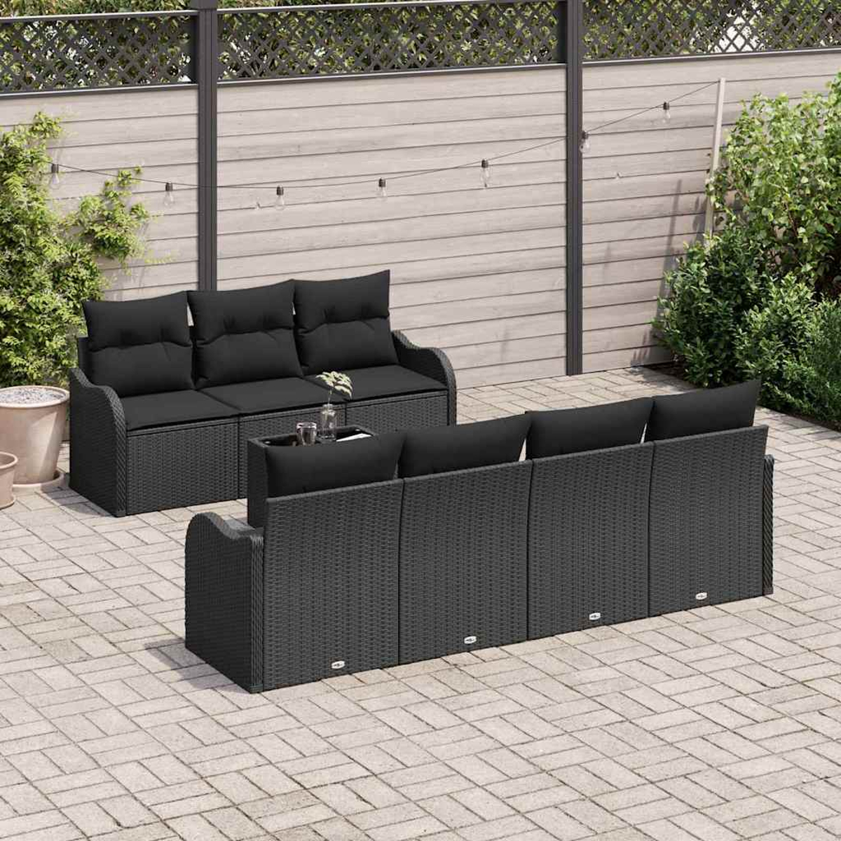 Zwei schwarze Korbsessel auf einer Terrasse mit Holzzaun im Hintergrund. Pflanzen umgeben die Terrasse. Lichterketten sind oben.