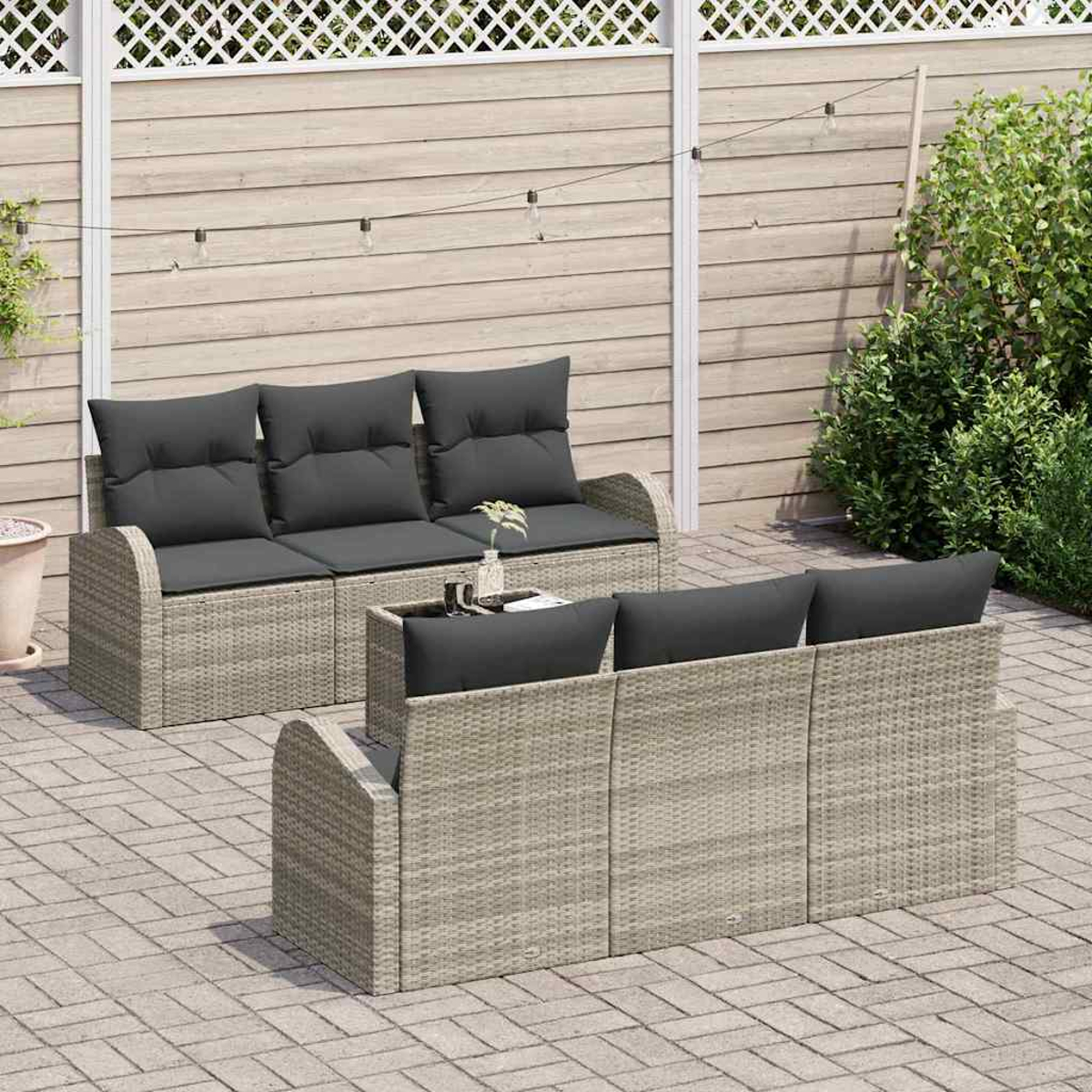 Outdoor-Rattansofas mit dunklen Kissen auf einer Ziegelterrasse. Im Hintergrund ein Holzzaun und grüne Büsche.