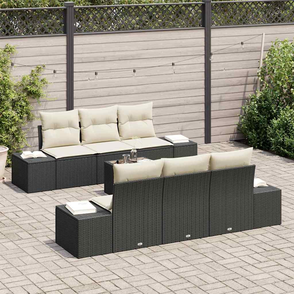 Juego de muebles de patio. Sofás de tejido gris oscuro con cojines crema. Fondo: valla de madera, plantas y luces de cadena.