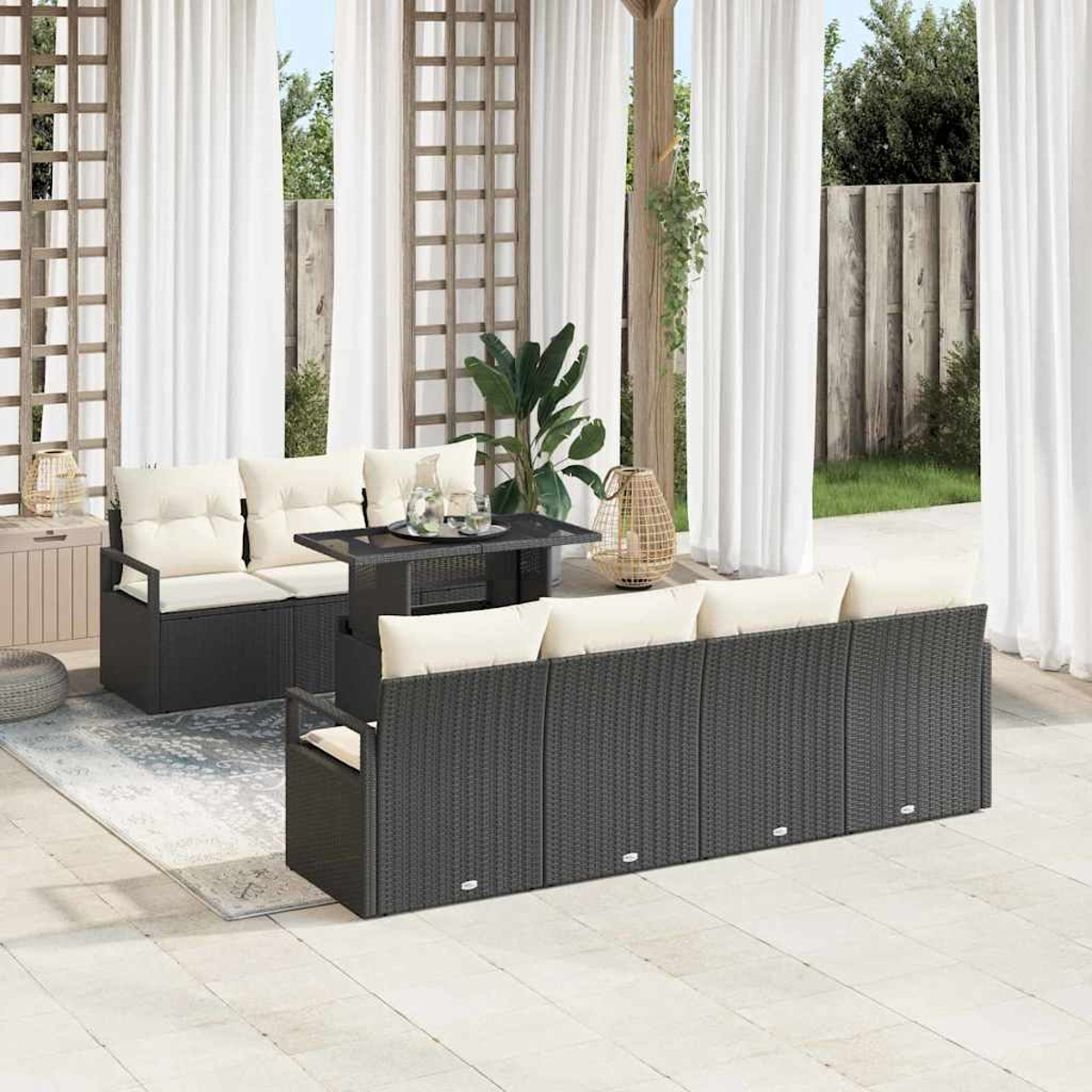 Terrassenset mit schwarzen Korbsesseln, cremefarbenen Kissen, einem schwarzen Tisch und weißen Vorhängen. Pflanzen und ein Holzzaun sind sichtbar.