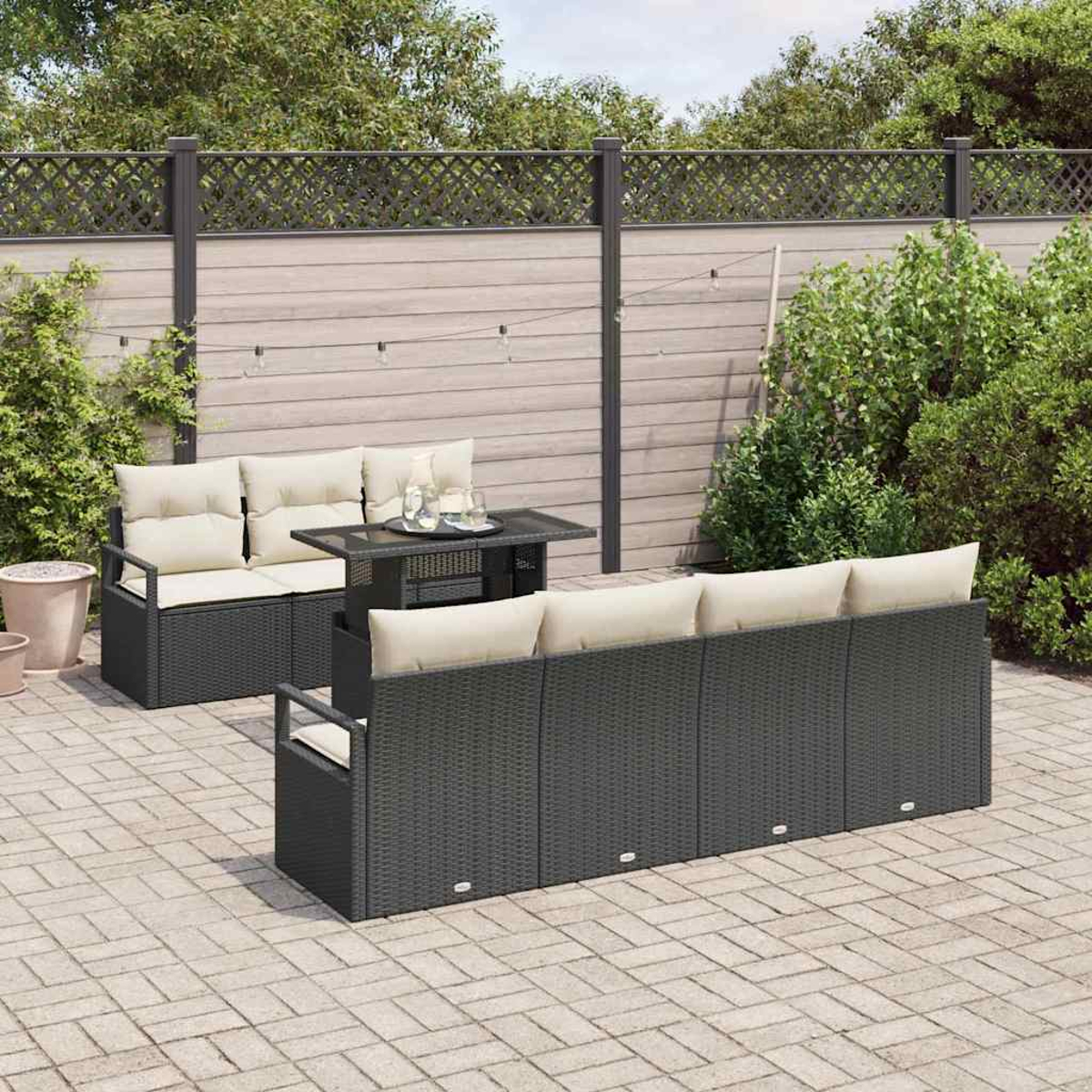 Garten-Terrassenset mit schwarzen Korbsesseln, beigen Kissen und einem Tisch mit Glasplatte. Ein Holzzaun und grüne Büsche im Hintergrund.