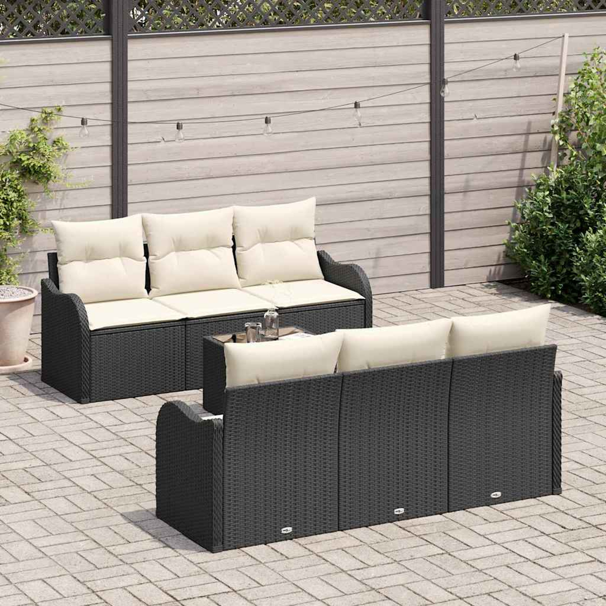 Juego de muebles de patio al aire libre: dos sofás de mimbre gris oscuro con cojines crema, sobre un patio de ladrillos. Fondo: valla de madera y plantas.