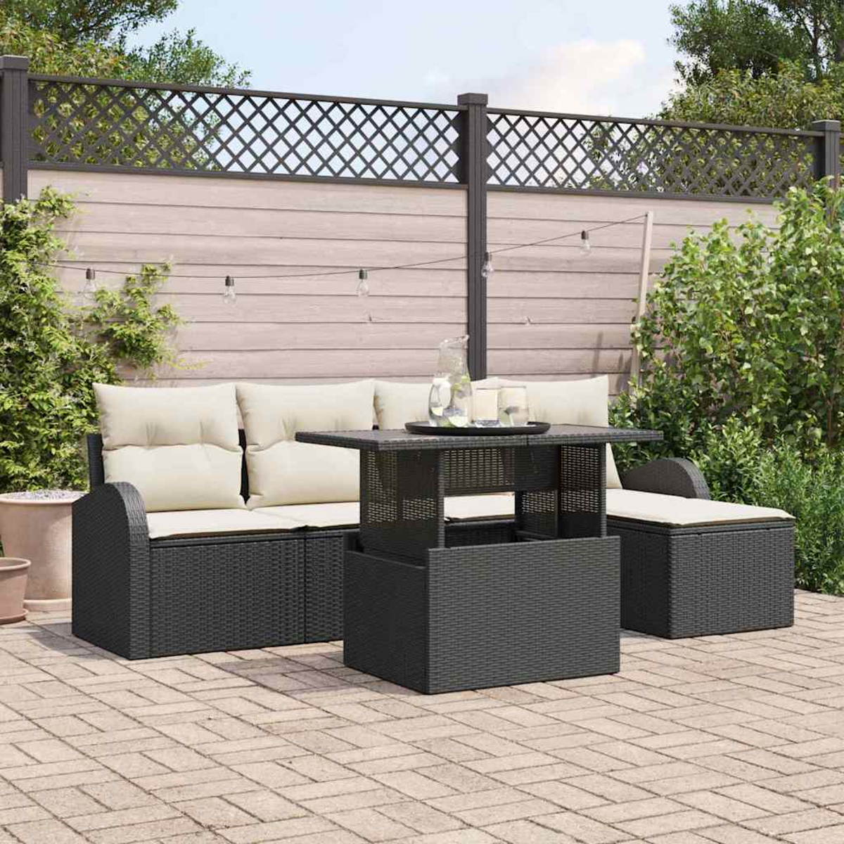 Garten-Terrassenset mit schwarzen Korbmöbeln, beigen Kissen und einem ansteigenden Tisch. Im Hintergrund befinden sich ein Zaun und Grün.