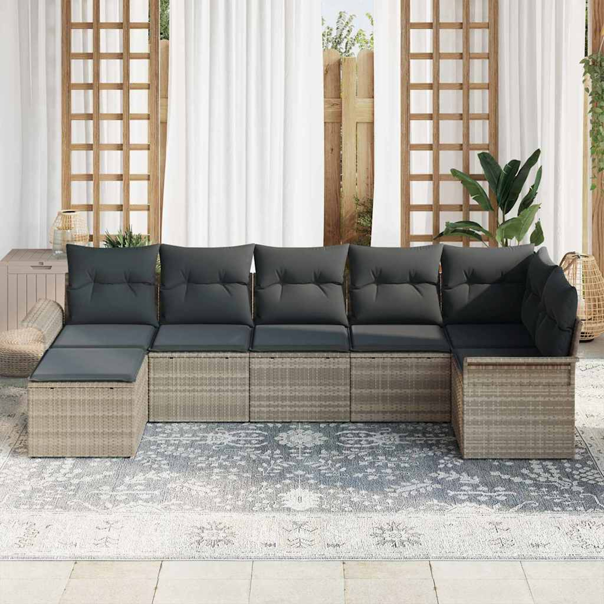 Ein Ecksofa im Freien mit dunkelgrauen Kissen steht auf einem gemusterten Teppich. Weiße Vorhänge und ein Holzzaun sind im Hintergrund.
