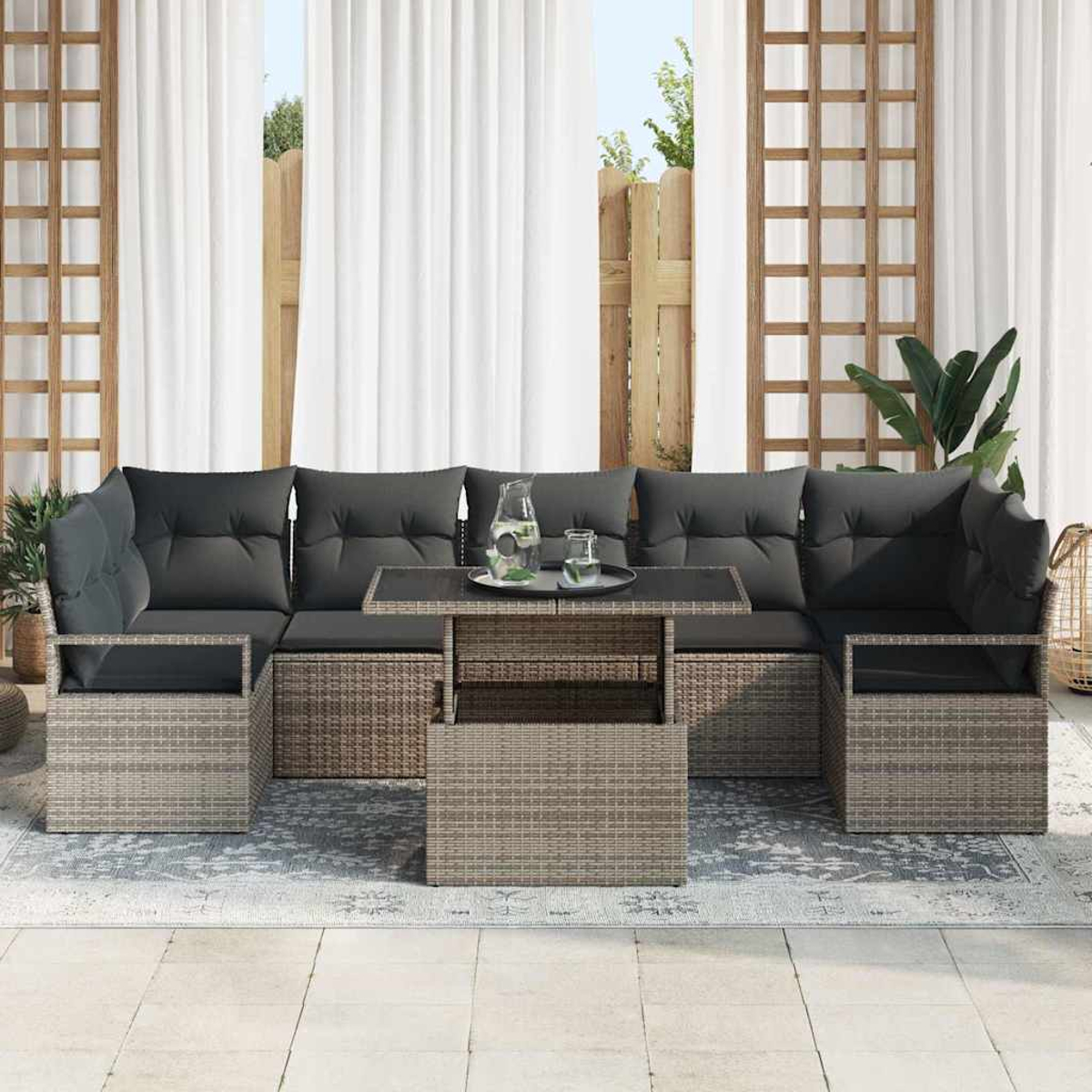 Juego de sofás de mimbre para exteriores con cojines grises y una mesa con tapa de cristal. Cortinas blancas y una valla de madera al fondo.