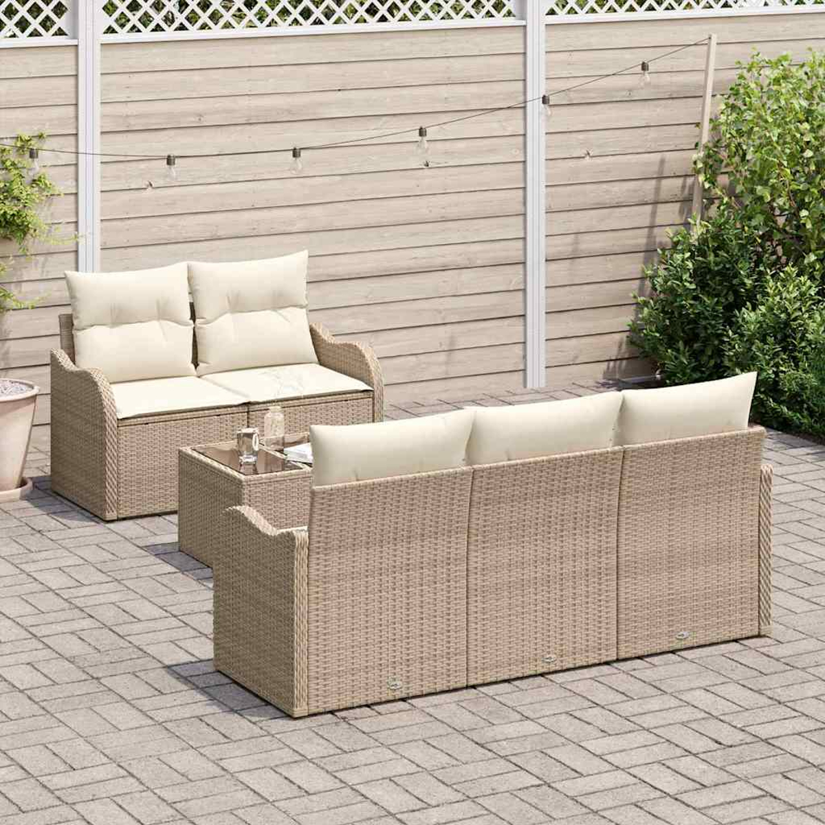 Outdoor-Rattanmöbel-Set mit beigen Kissen und einem Glastisch. Das Set steht auf einer Steinterrasse mit Holzzaun und grünen Pflanzen.