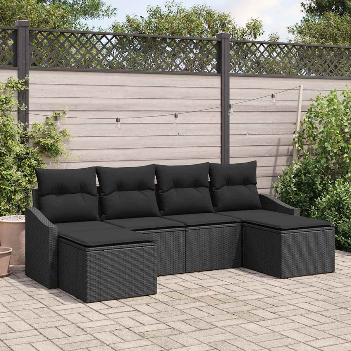 Ein schwarzes Korbsessel-Ecksofa steht auf einer Backstein-Terrasse. Ein Holzzaun mit Gitteraufsatz befindet sich hinter dem Sofa.