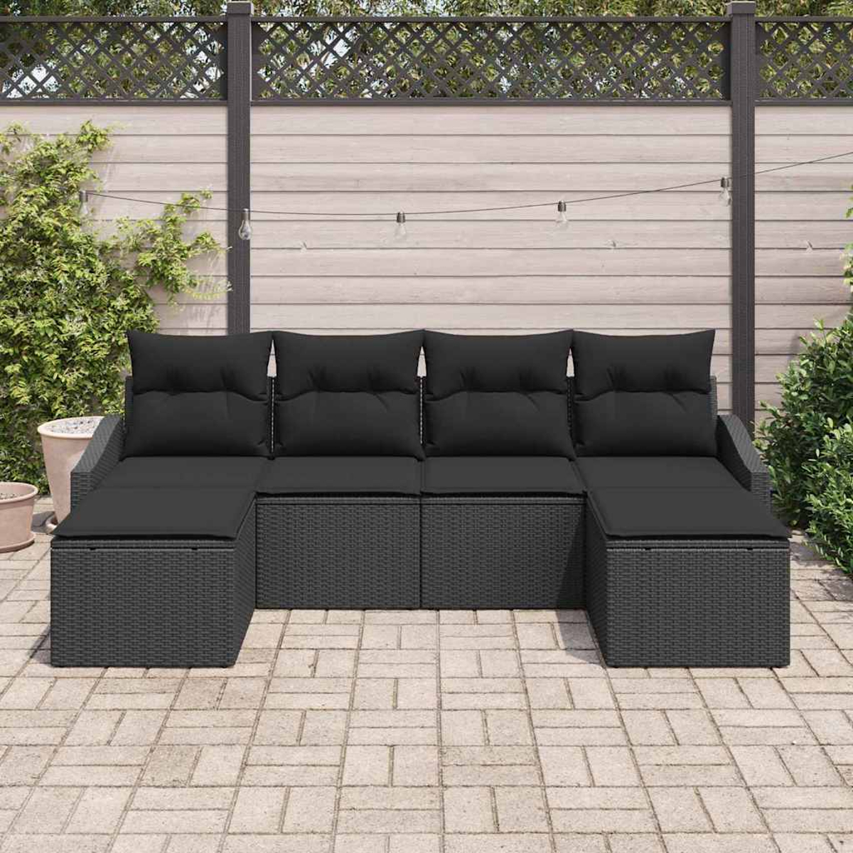 Ein schwarzes Outdoor-Ecksofa steht auf einer Steinterrasse. Im Hintergrund befinden sich ein Holzzaun mit Grün und Lichterketten.