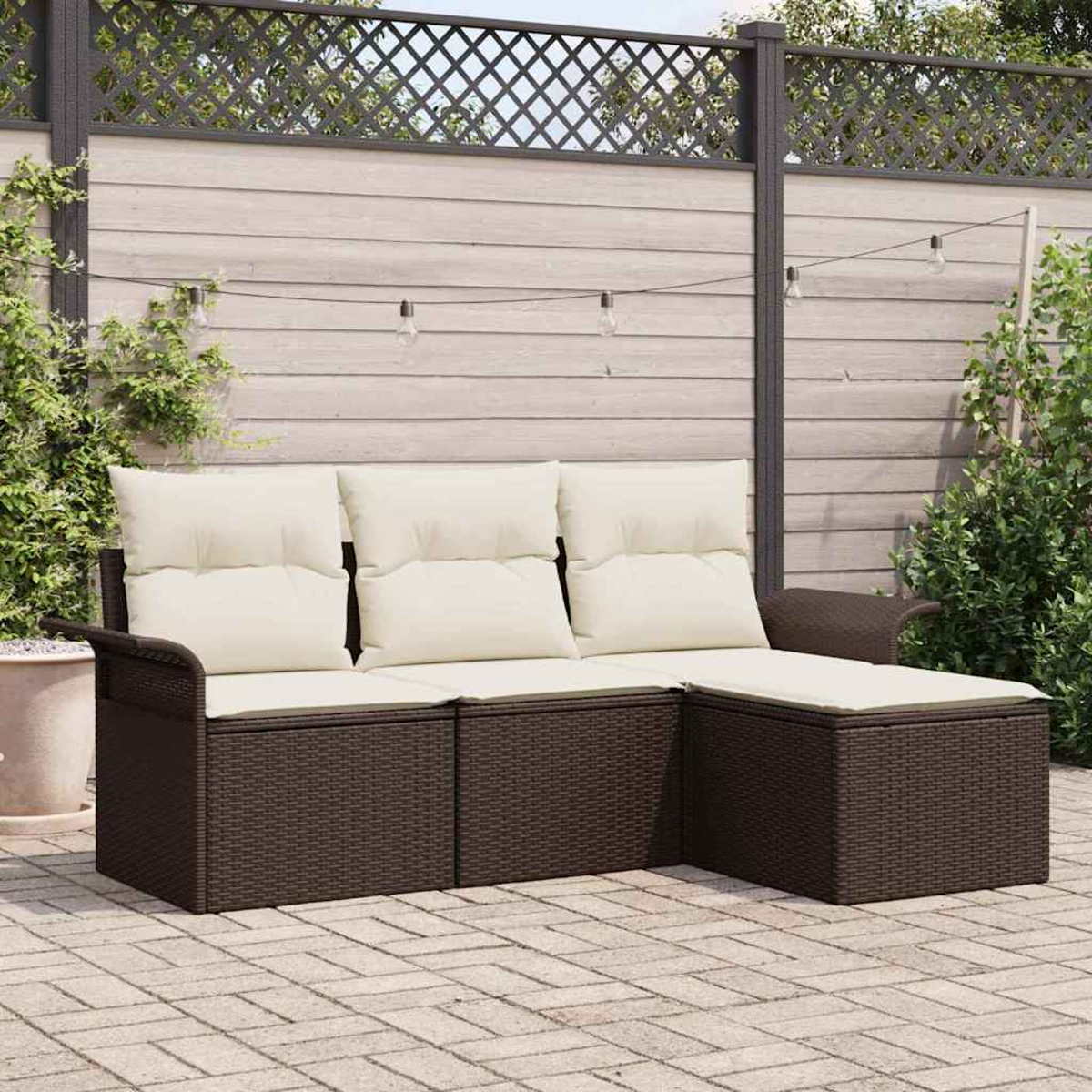 Ein braunes, aus Weide geflochtenes Ecksofa mit cremefarbenen Kissen steht auf einer Backstein-Terrasse. Dahinter ist ein Holzzaun mit Lichterketten.