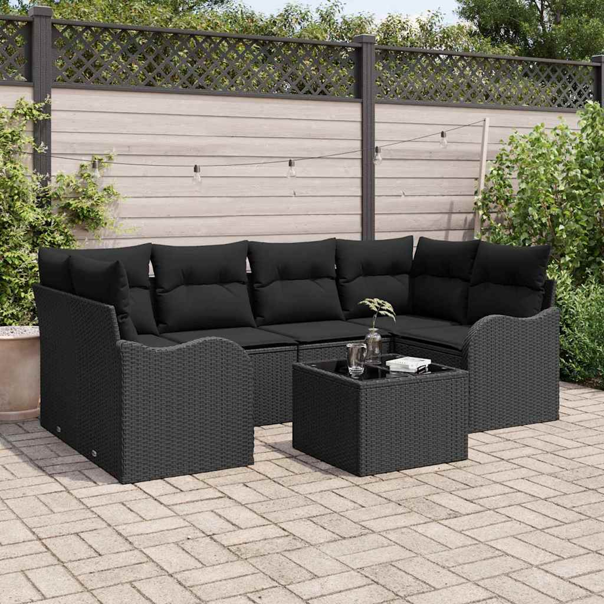 Garten-Sitzgruppe mit schwarzen Kissen, einem kleinen Tisch, auf einer Backstein-Terrasse. Ein Holzzaun mit Grün im Hintergrund.