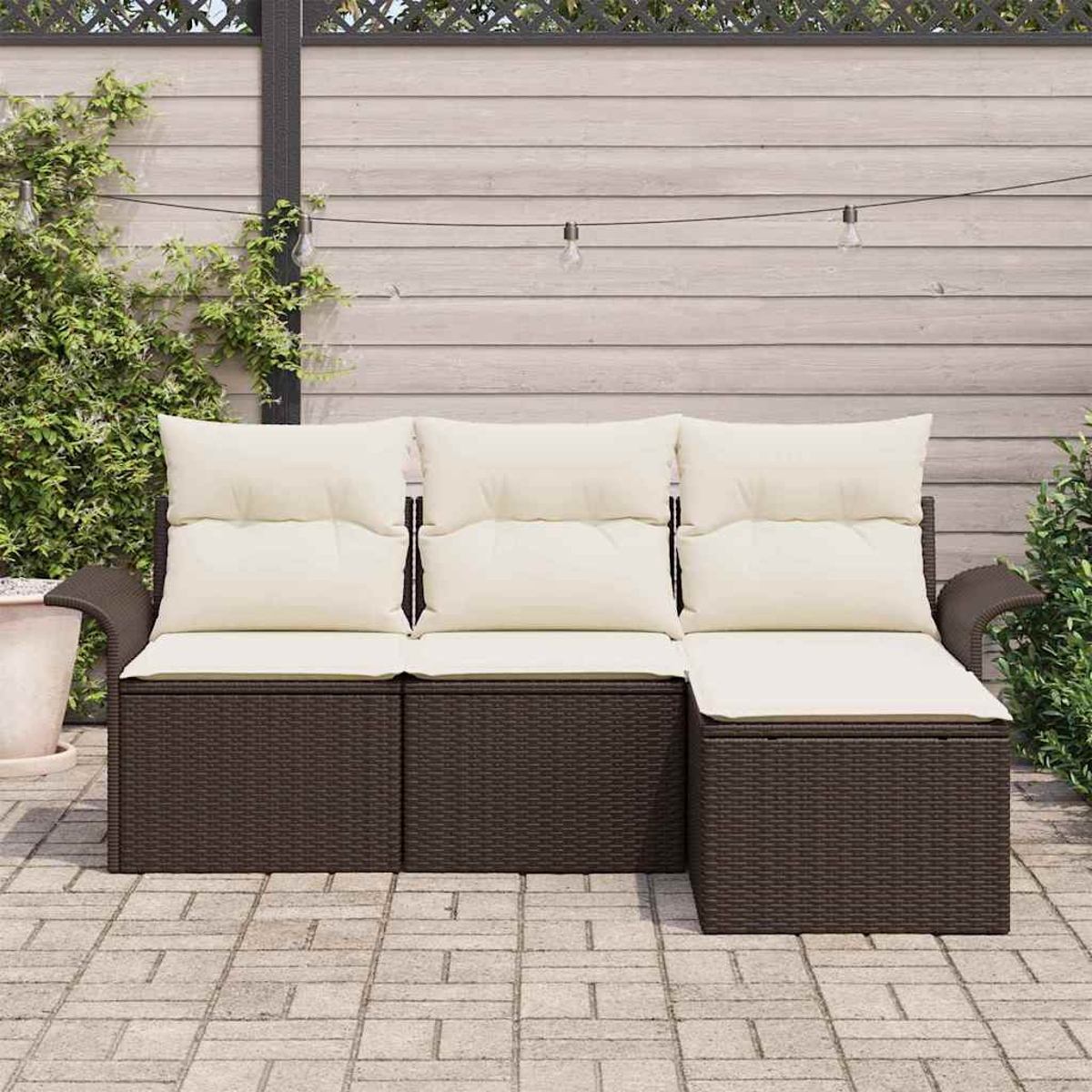 Ein braunes Outdoor-Sofa aus Korbgeflecht mit cremefarbenen Kissen steht auf einer Backstein-Terrasse. Im Hintergrund befinden sich ein Holzzaun und Pflanzen.