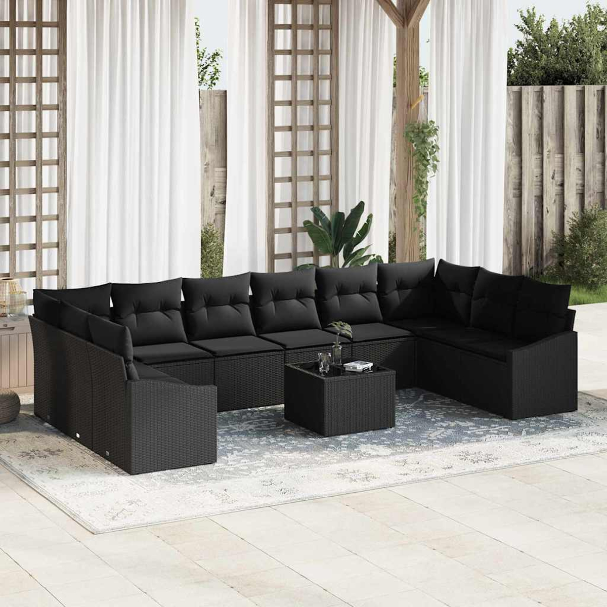 Schwarzes Ecksofa mit Kissen und einem Couchtisch auf einem gemusterten Teppich unter einer Pergola mit weißen Vorhängen.
