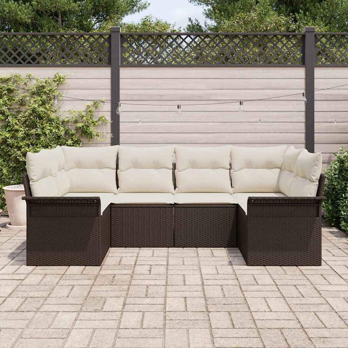 Ein braunes Outdoor-Ecksofa aus Korbgeflecht mit cremefarbenen Kissen steht auf einer Steinterrasse. Der Hintergrund umfasst einen Zaun und Grün.