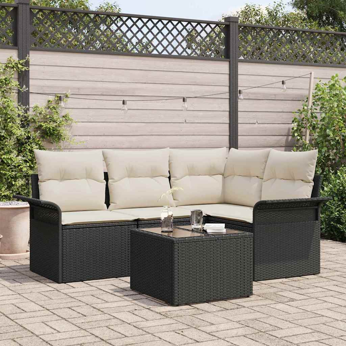 Schwarzes Korbmöbel-Set mit cremefarbenen Kissen und einem Tisch mit Glasplatte auf einer gepflasterten Terrasse.