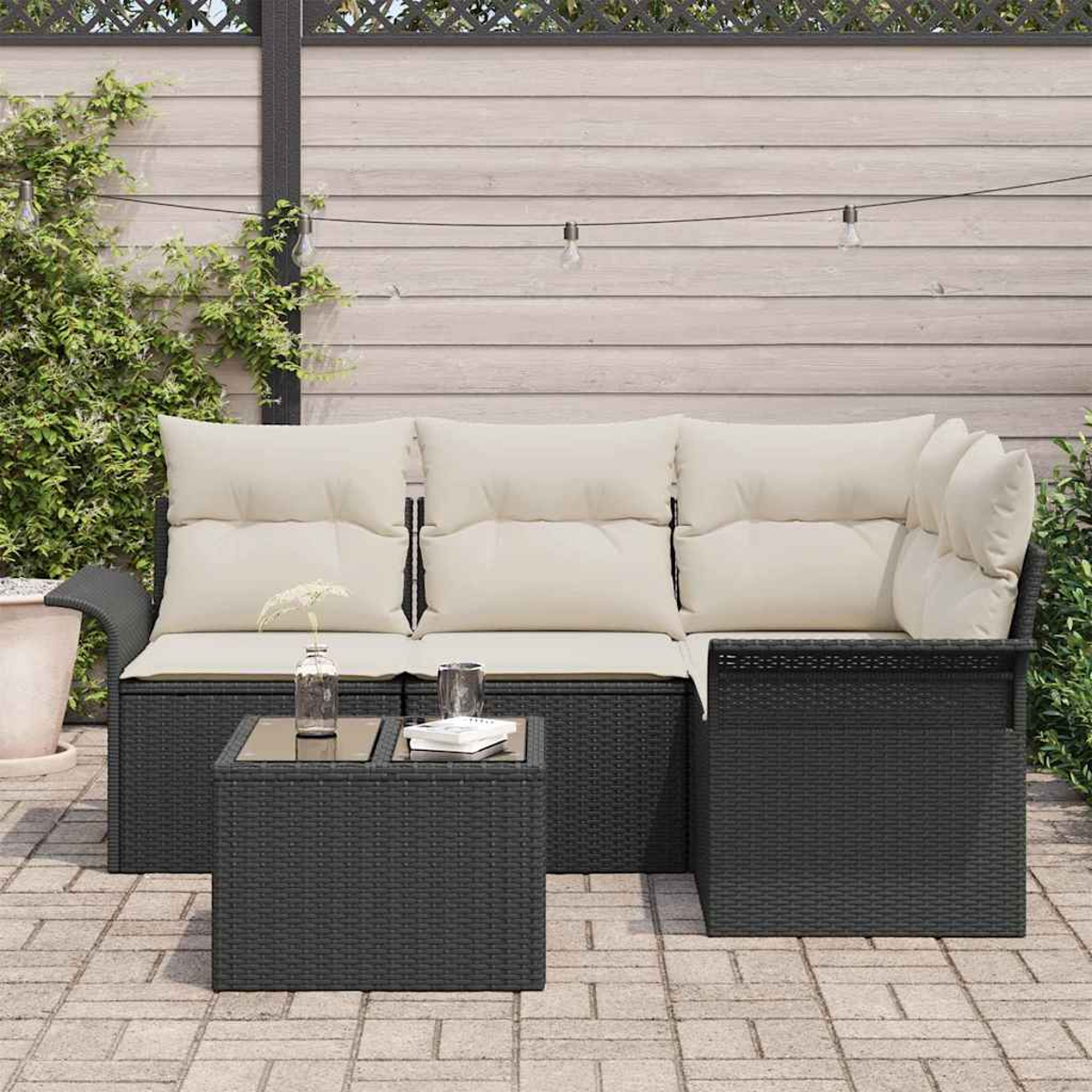 Gartenmöbel-Set mit schwarzem Korbsessel, cremefarbenen Kissen und einem kleinen Tisch. Ein Holzzaun und Pflanzen vervollständigen die Szene.