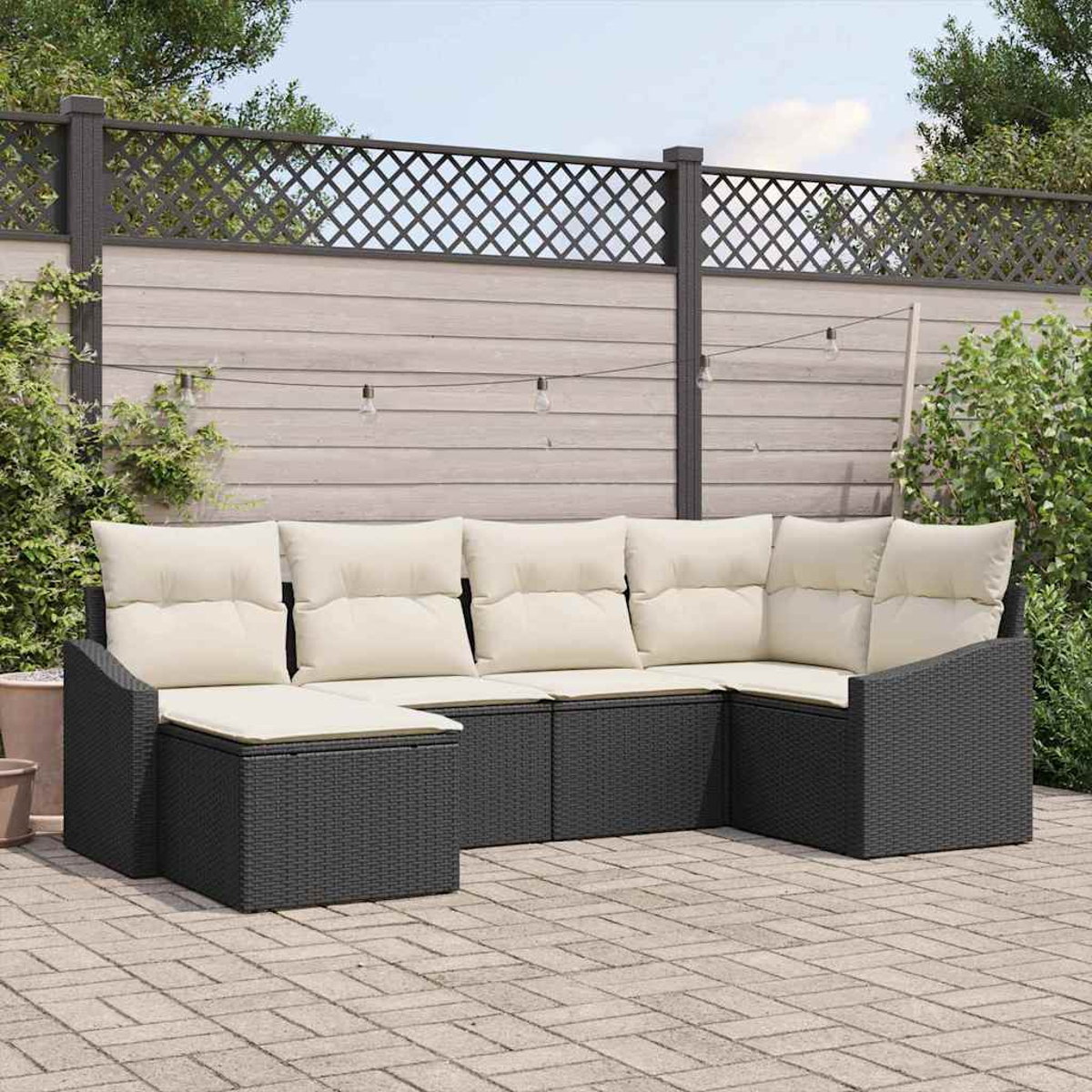 Dunkelgraues Outdoor-Sofa aus Korbgeflecht mit cremefarbenen Kissen steht auf einer Ziegelterrasse. Im Hintergrund ein Holzzaun mit Lichterketten.