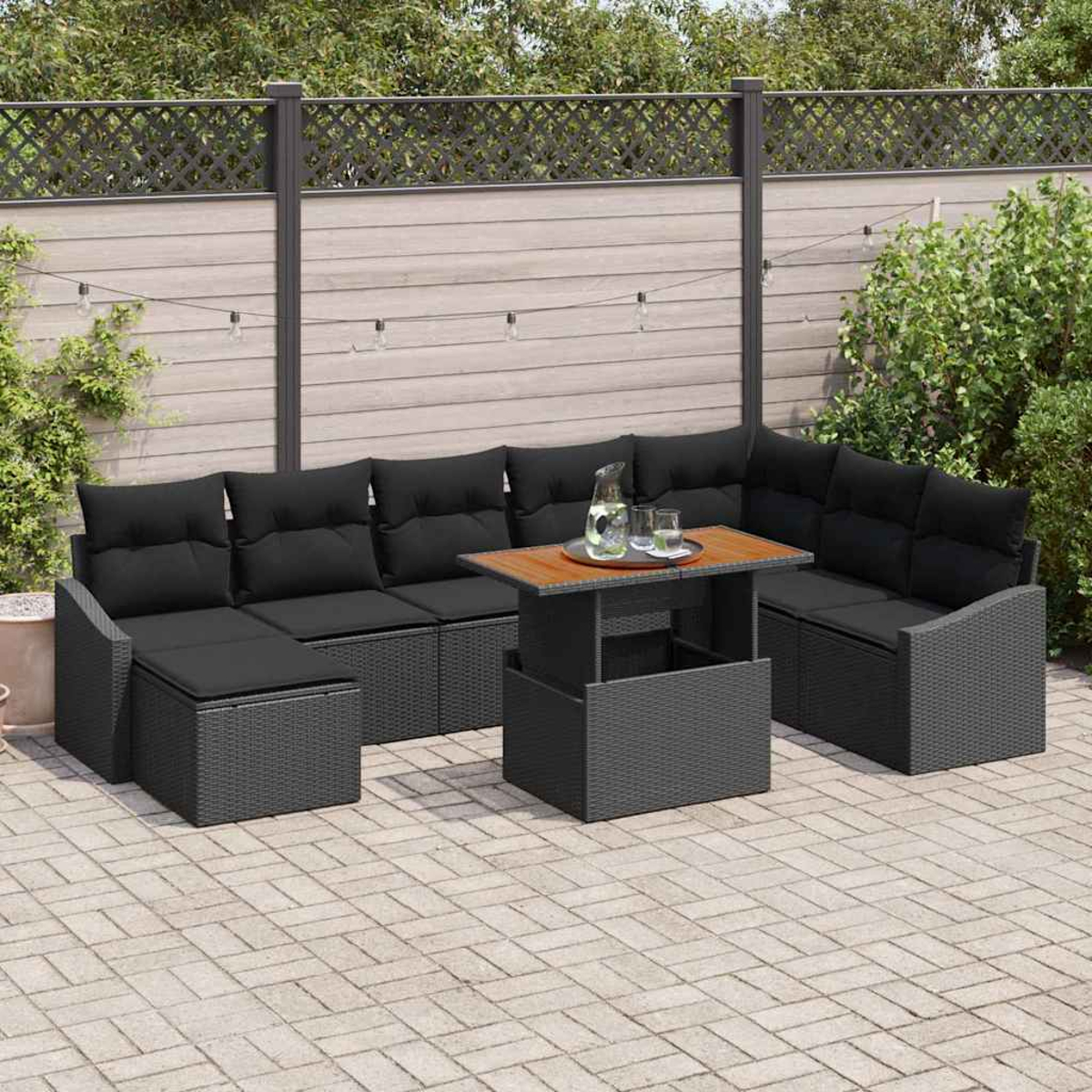 Gartenmöbel-Set mit schwarzen Kissen, einem Holztisch und Gläsern auf einer Terrasse. Im Hintergrund sind ein Zaun und Pflanzen zu sehen.