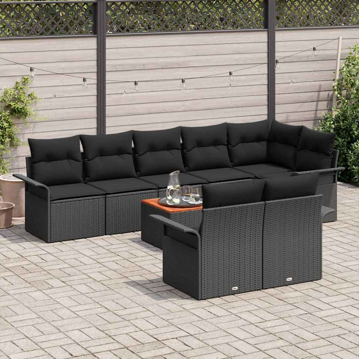 Juego de patio al aire libre con cojines negros y mimbre gris. Incluye una mesa con vasos y una jarra. Valla y plantas de fondo.