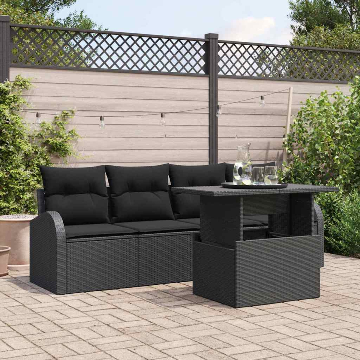 Schwarzes Korbmöbel-Set mit Sofa und Tisch im Garten. Holzzaun und Pflanzen im Hintergrund. Getränke auf dem Tisch.