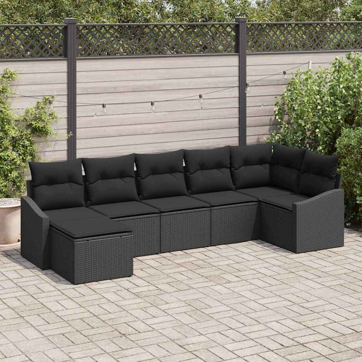 Ein schwarzes Ecksofa mit Kissen steht auf einer Terrasse. Im Hintergrund sind ein Holzzaun und Lichterketten zu sehen.