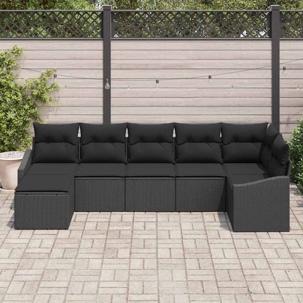 Ein schwarzes Korbsessel-Sofa mit schwarzen Kissen steht auf einer Terrasse. Ein Holzzaun mit Grün und Lichterketten ist im Hintergrund.