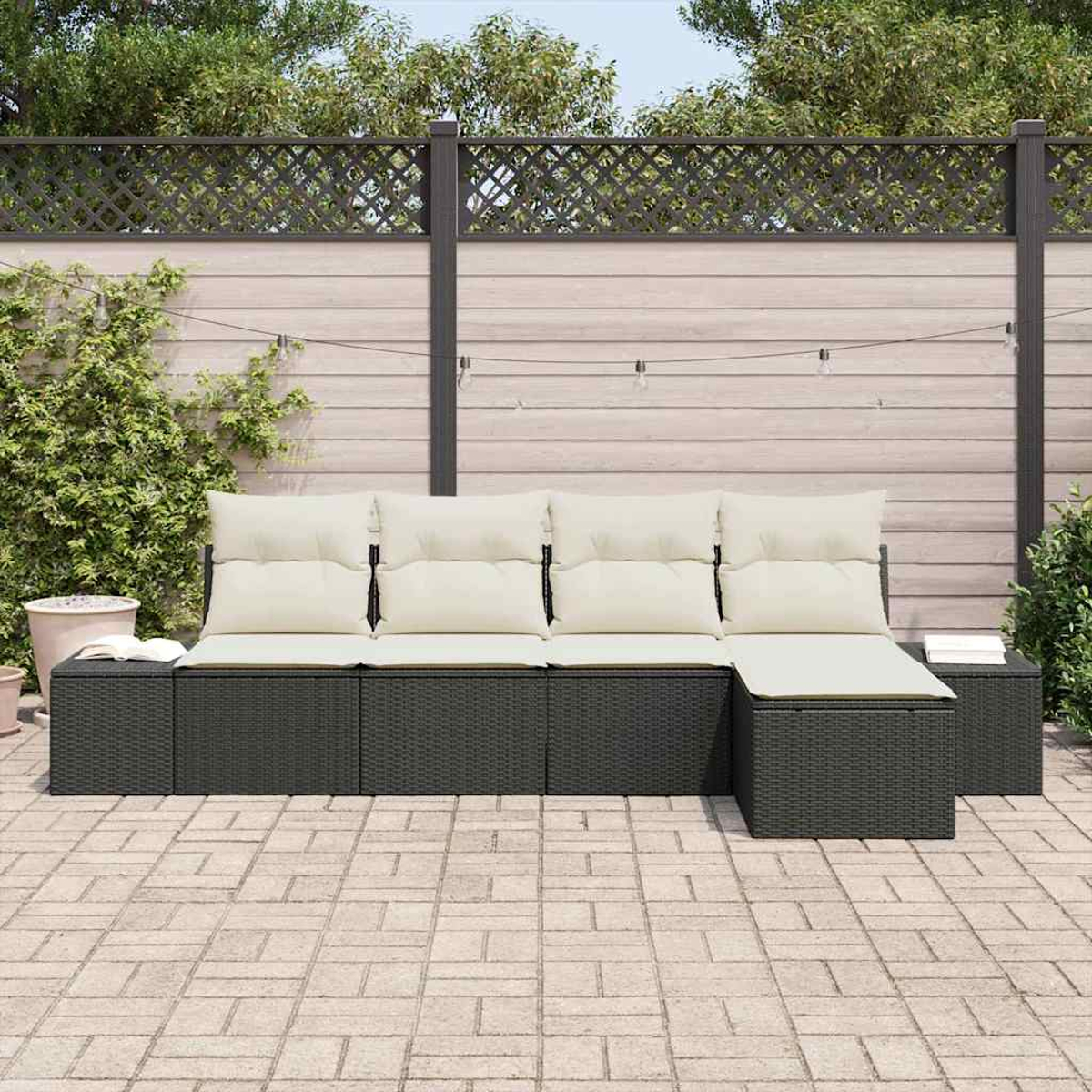 Outdoor-Ecksofa mit cremefarbenen Kissen. Dahinter befindet sich ein Zaun mit Kletterpflanzen und Lichterketten. Ziegelterrasse im Vordergrund.