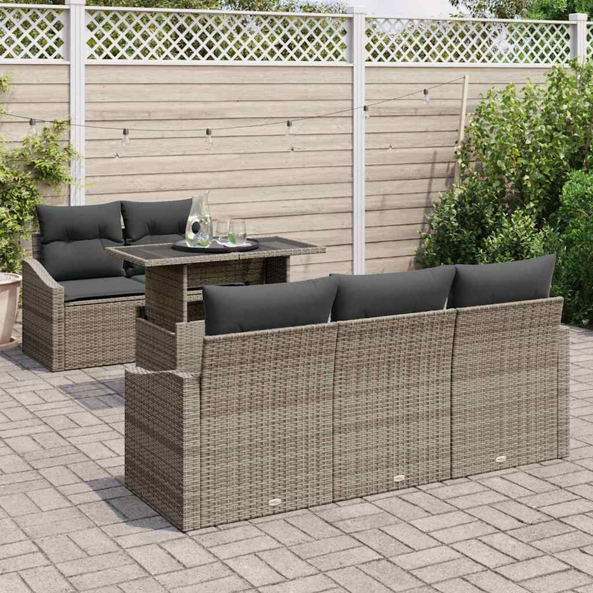 Gartenmöbel-Set aus Korbgeflecht mit grauen Kissen und einem Tisch. Auf einer Ziegelterrasse, mit Gitterzaun und Grün im Hintergrund.