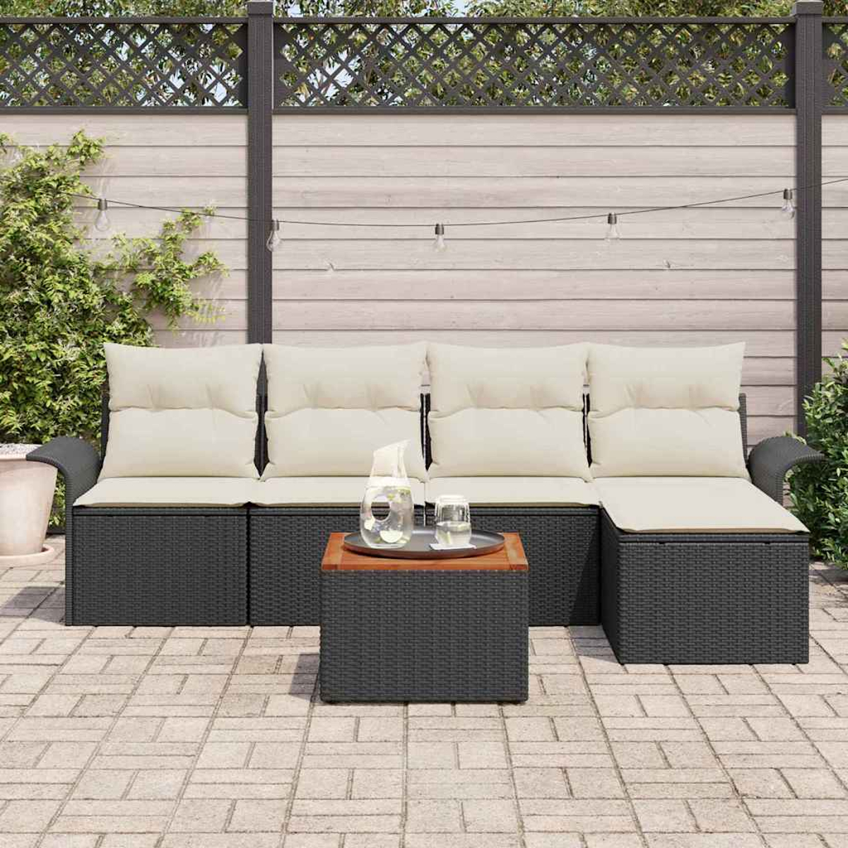 Outdoor-Ecksofa mit cremefarbenen Kissen und einem kleinen Tisch. Hintergrund ist ein Zaun, Lichterketten und Grün.