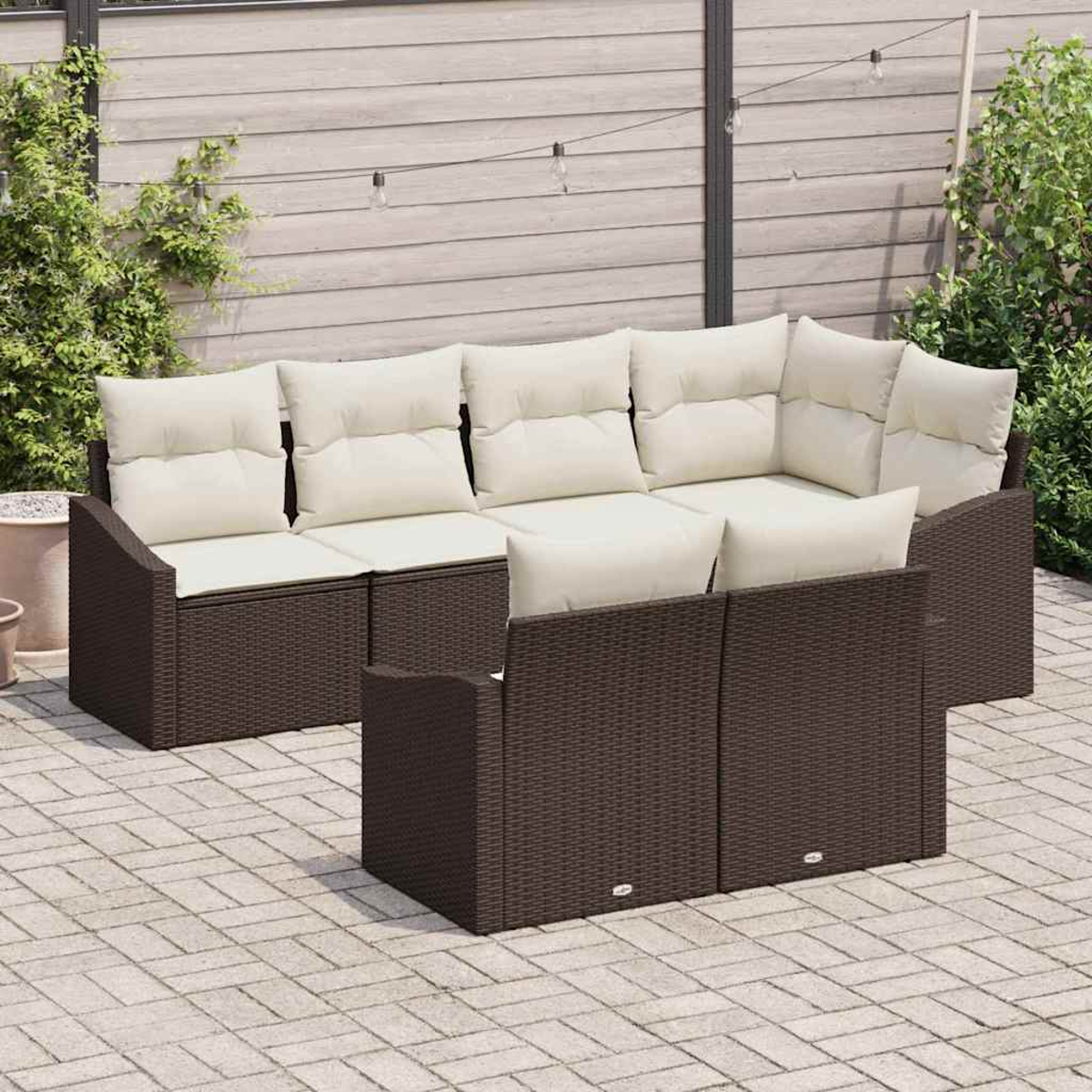 Braunes Outdoor-Korbmöbel-Set mit cremefarbenen Kissen. Auf einer Terrasse mit Pflanzen und Lichterketten im Hintergrund.