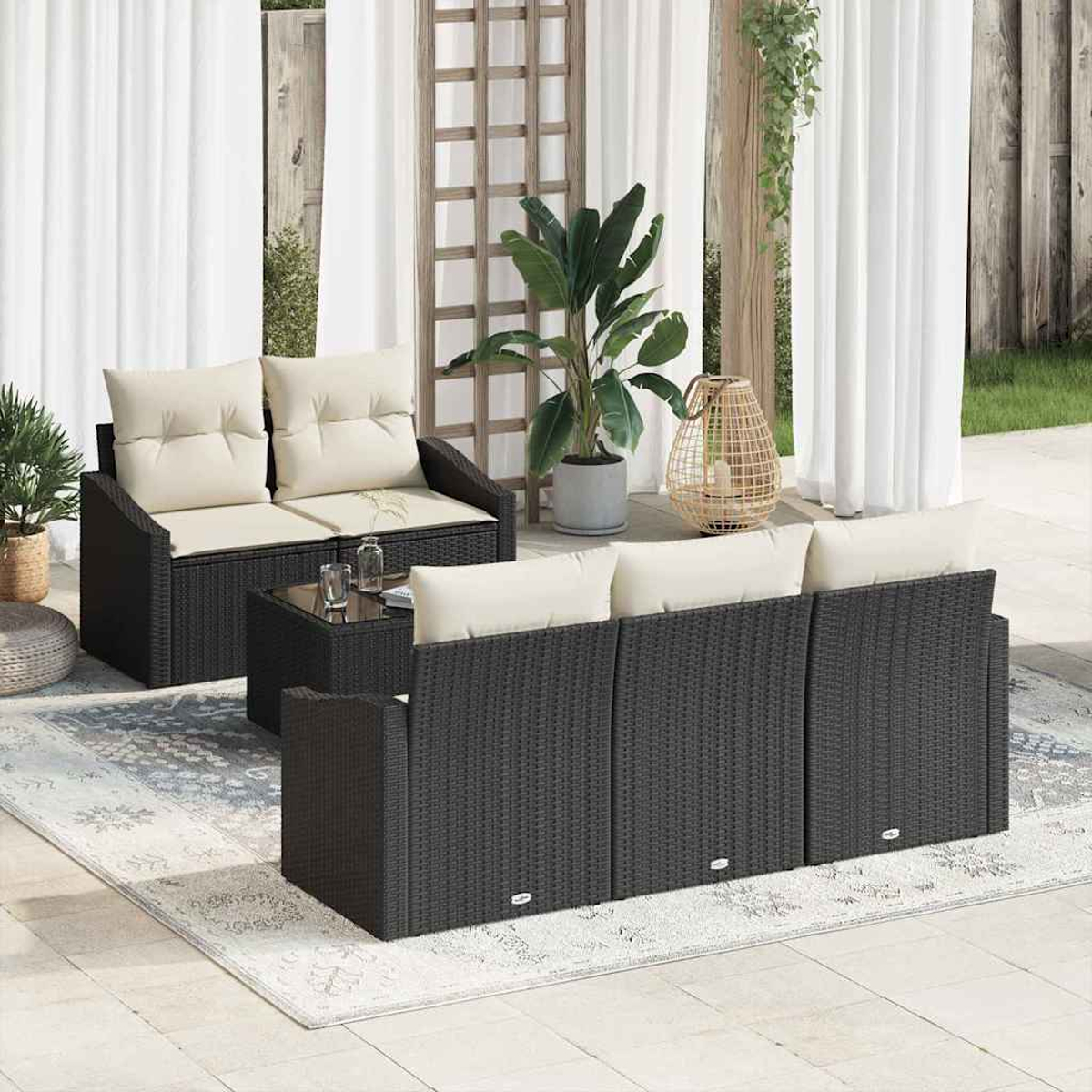 Gartenmöbel-Set mit schwarzem Korbsessel, Zweisitzer und Tisch mit Glasplatte und cremefarbenen Kissen. Hintergrund Pflanzen und weiße Vorhänge.