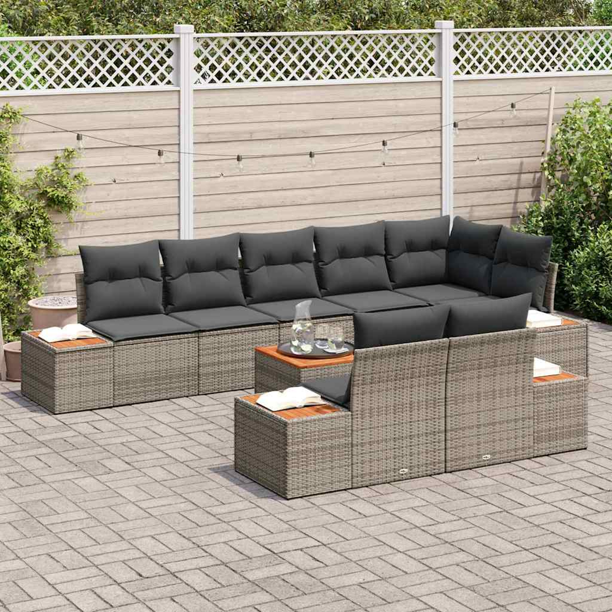 Garten-Lounge-Set: graues Korbmöbel-Ecksofa mit dunkelgrauen Kissen und Holztischen. Hintergrund: Zaun und Pflanzen.