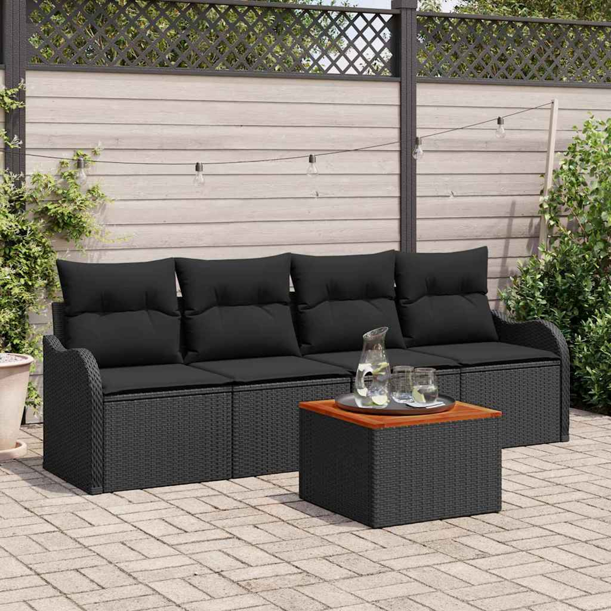 Schwarzes Outdoor-Korbmöbel-Set mit Kissen und Tisch mit Gläsern und Krug. Im Hintergrund Zaun und Pflanzen.
