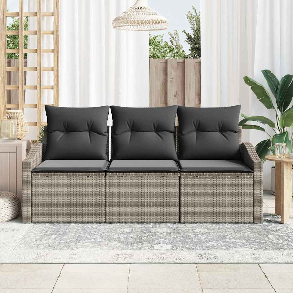 Ein Rattansofa mit dunkelgrauen Kissen steht auf einem Teppich. Ein weißer Vorhang und eine Hängelampe sind sichtbar. Rechts ist eine Pflanze.
