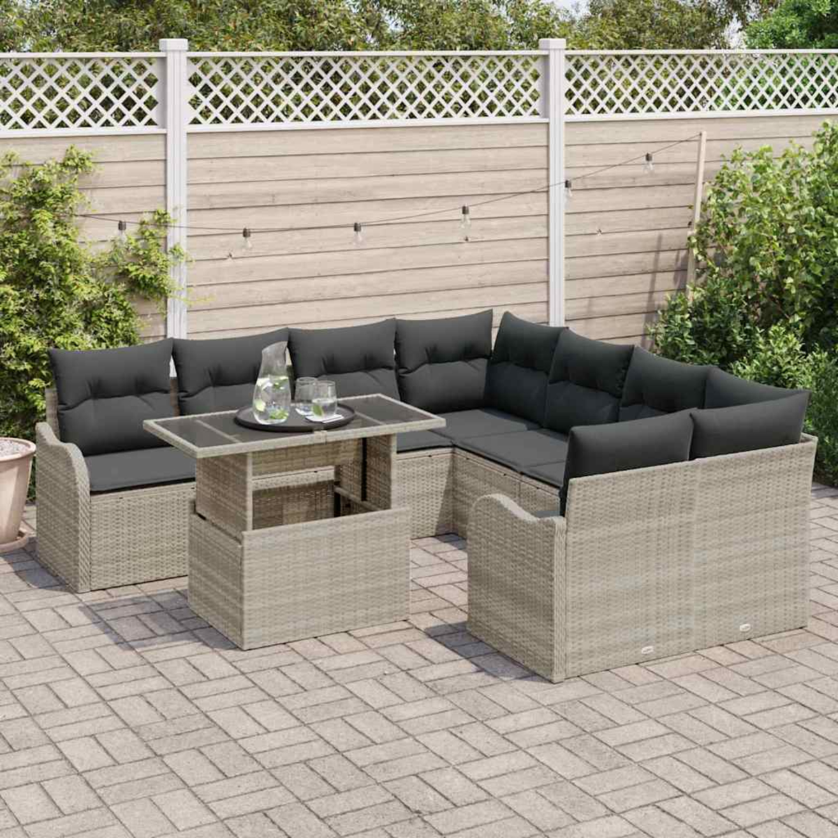 Ein graues Outdoor-Rattan-Ecksofa mit schwarzen Kissen, einem passenden Tisch mit Getränken und einem Holzzaun mit Grün.